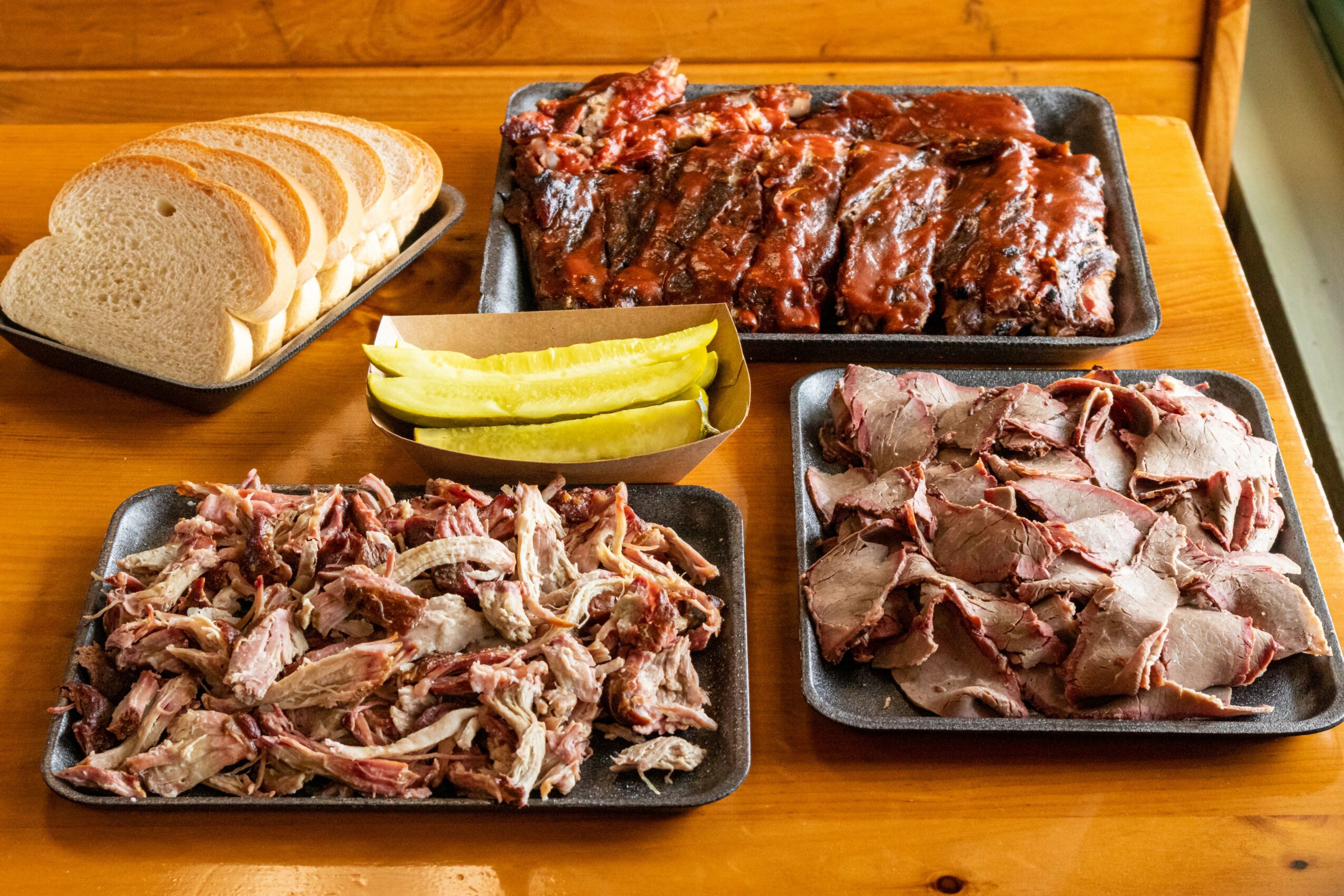 Slices of bread, raw ribs, chopped meat, and pickles on trays and a wooden table.
