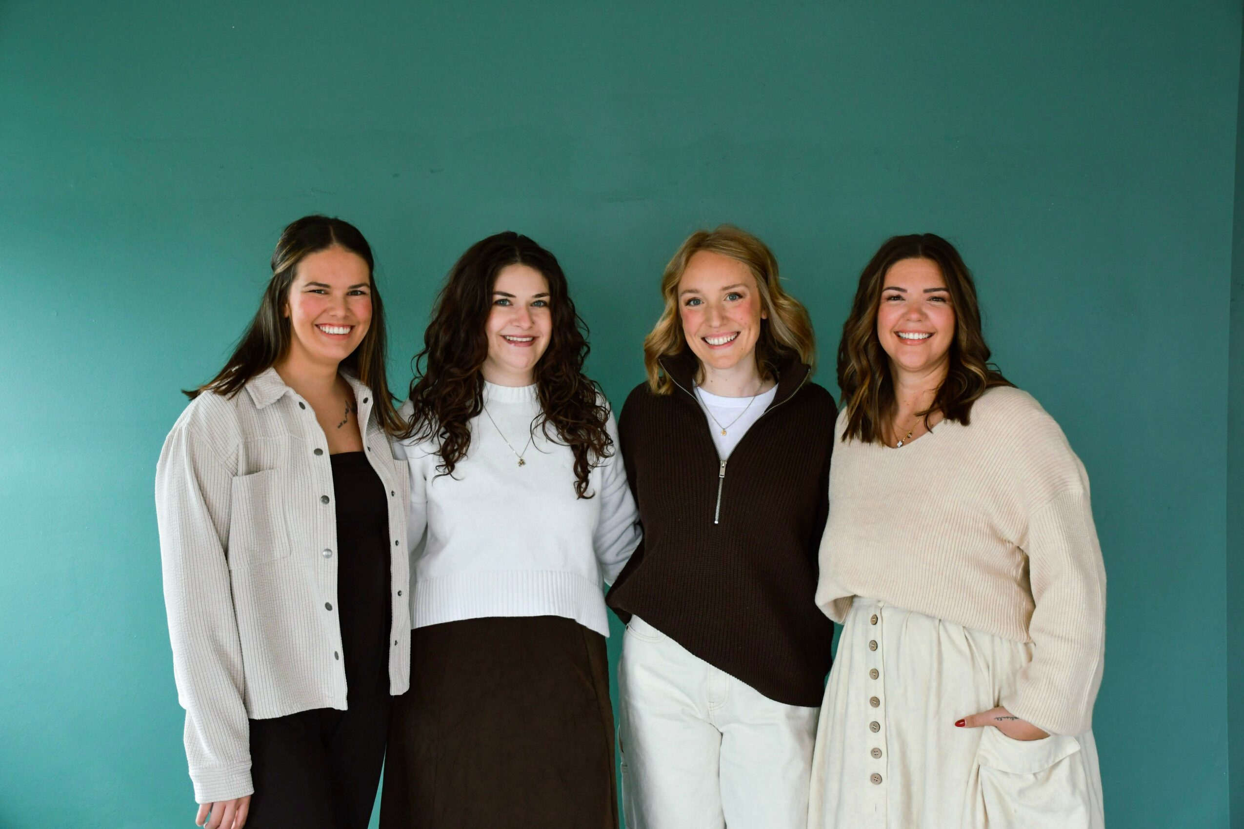 Four women standing together, smiling, against a teal background.