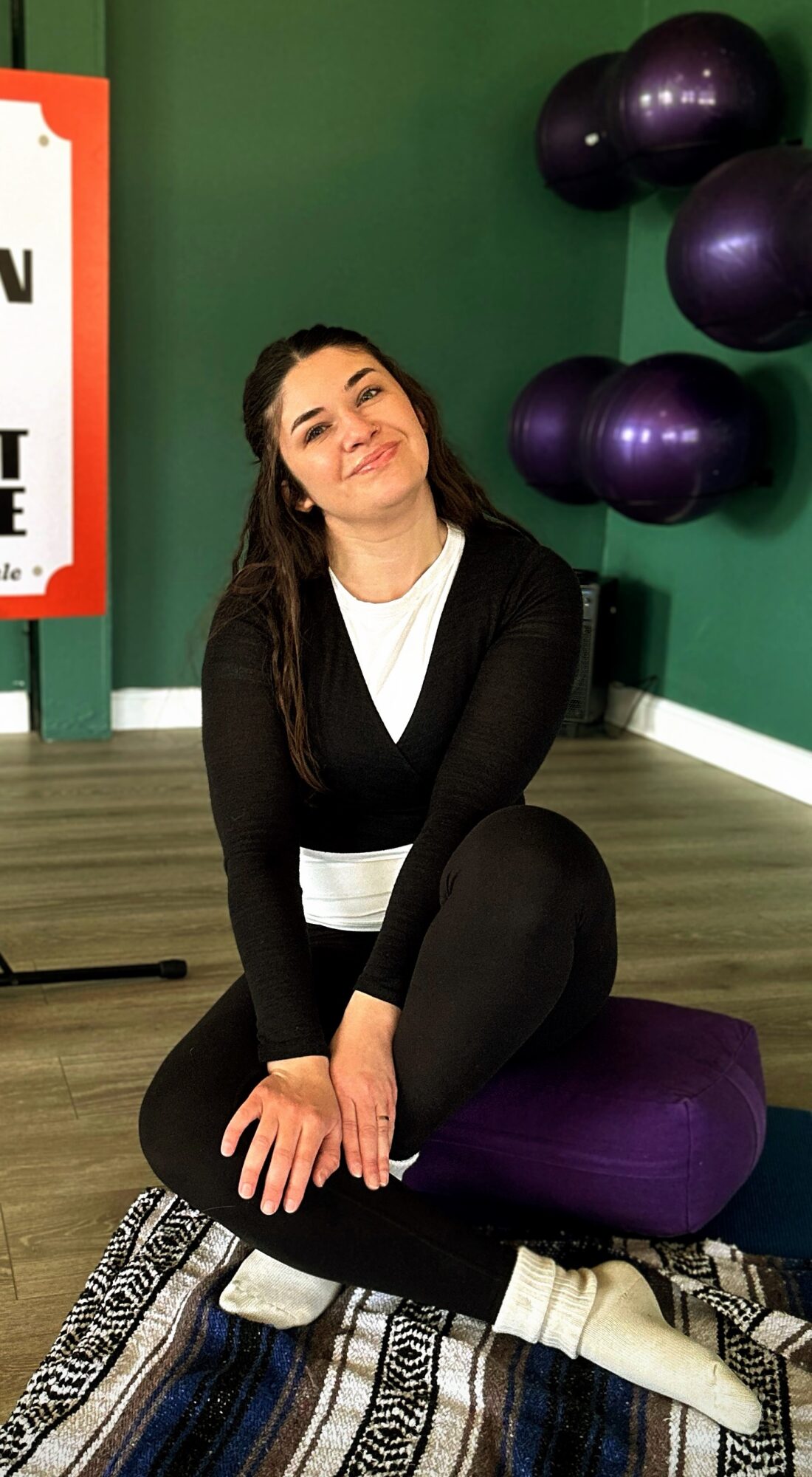 Woman sitting on a purple cushion, smiling, with black balloons on green wall background, and a patterned rug.