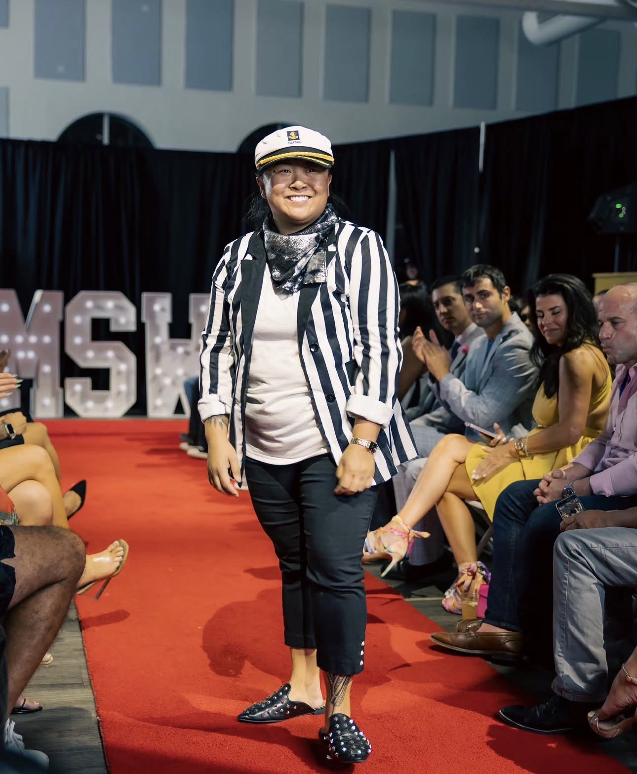 Woman walking on red carpet, smiling, wearing striped blazer, white shirt, black pants, and polka dot shoes, audience seated nearby.