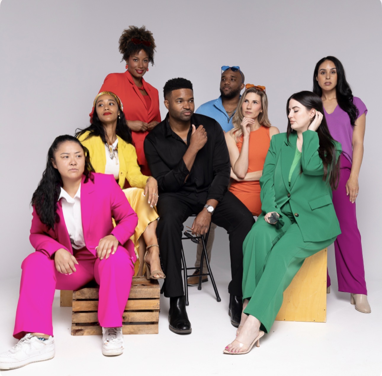 Group of nine diverse people in colorful outfits posing against a plain background.