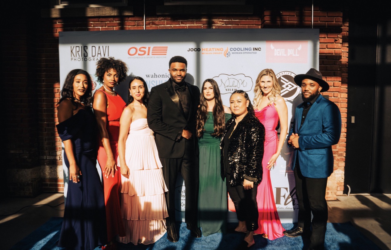 Group of eight people in formal attire standing outdoors in front of a backdrop with logos, sunlight casting shadows.