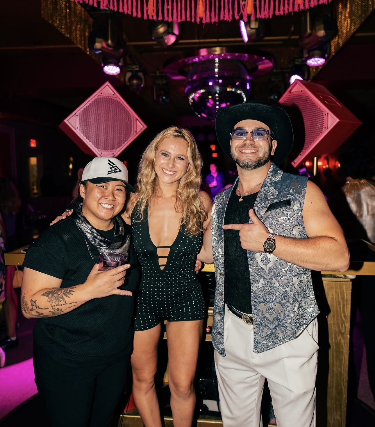 Three people smiling at a party with colorful lighting and large pink speakers in the background.