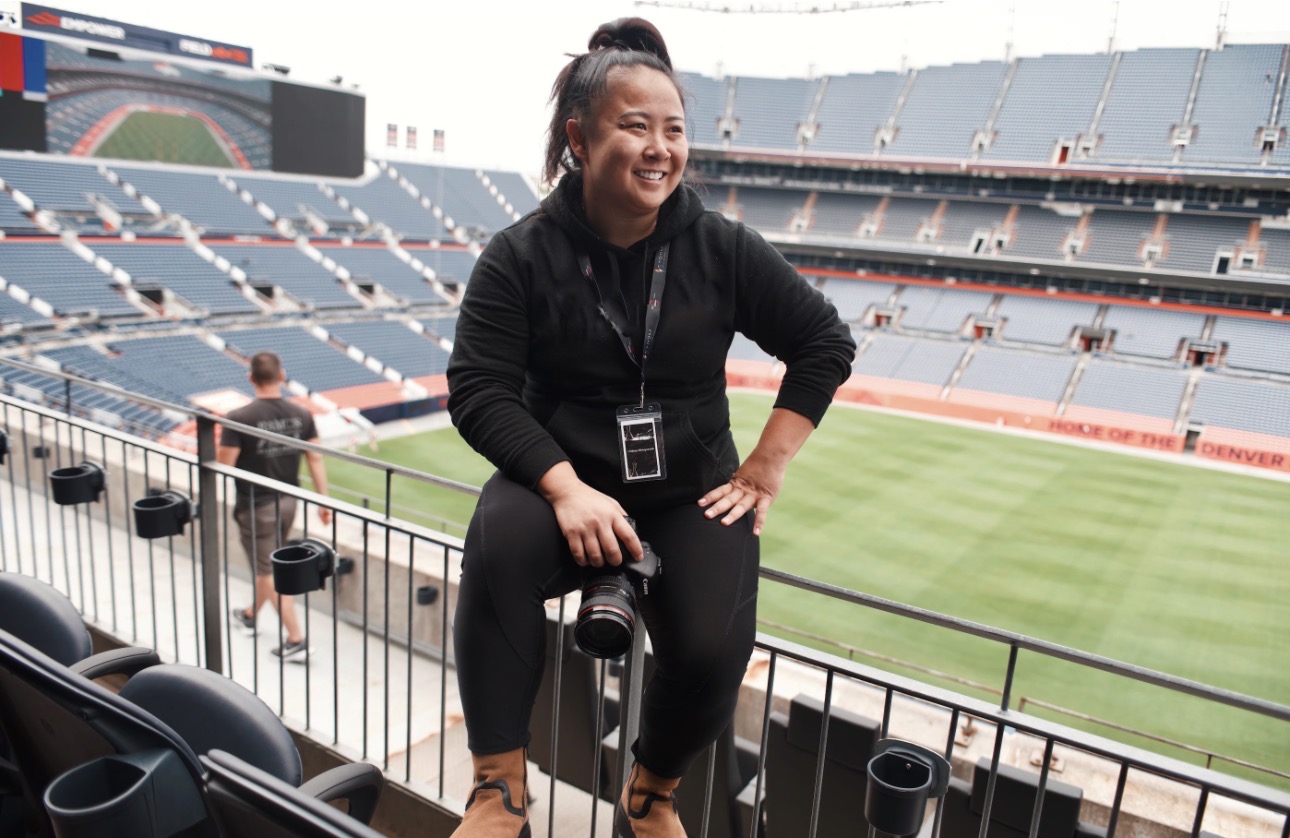 Smiling woman sitting on a railing in a stadium with empty seats and a field in the background.