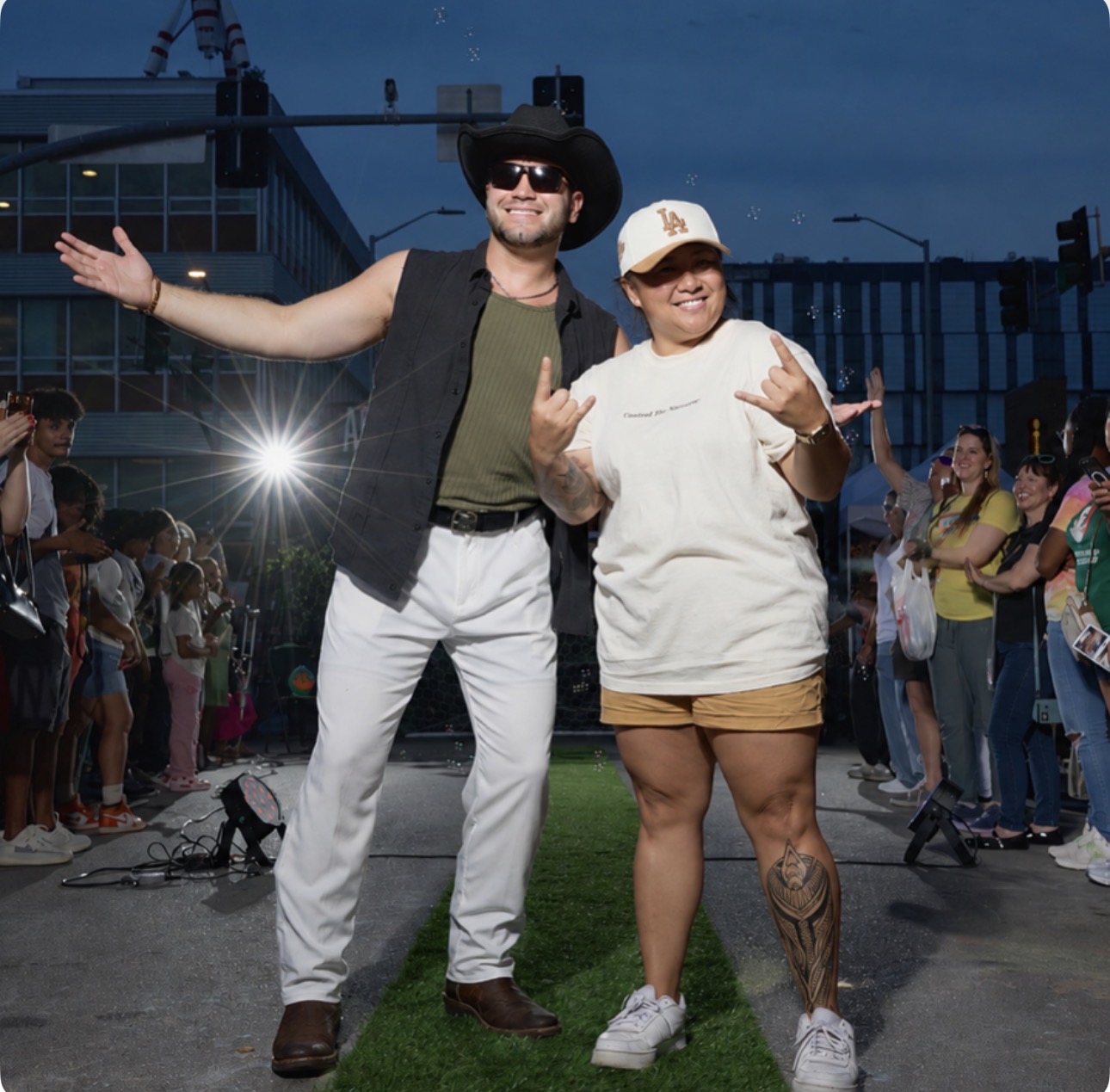 Two people posing outdoors with a crowd and city buildings in the background, one with arms outstretched, smiling.