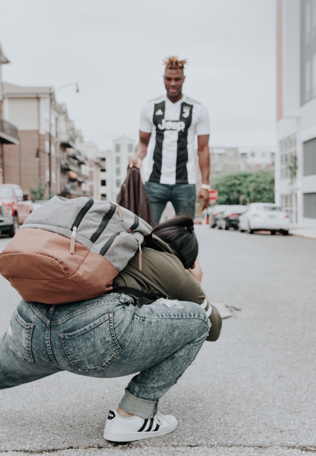 Person crouching on street with backpack, facing away, another person standing in background, urban setting, overcast sky.