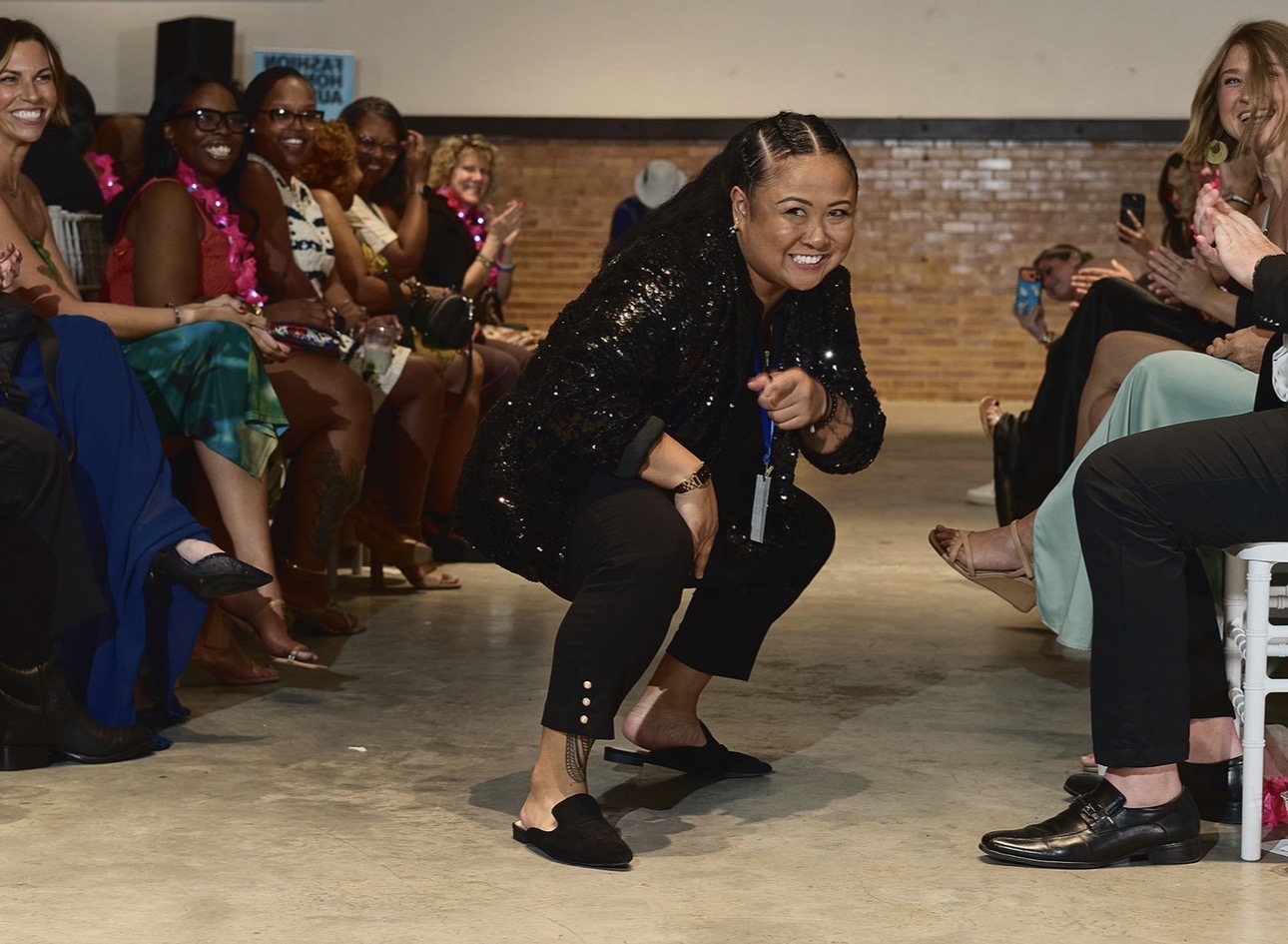 Woman crouching and smiling in a black outfit, surrounded by seated people in a room with a brick wall.