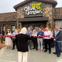 Group of people at ribbon-cutting event outside Olive Garden restaurant, with a woman speaking to the crowd.