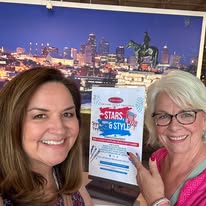 Two women smiling, one pointing at a sign, with a cityscape backdrop behind them.