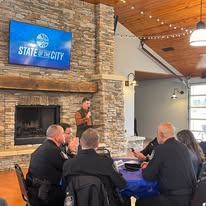 People sitting around a table in a room with a stone fireplace and a large screen displaying 'State of the City'.