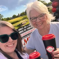 Two women smiling outdoors, one holding a coffee cup, with a sign and trees in the background.