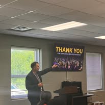Man in suit giving a presentation in a room with windows, a banner reading 'THANK YOU,' and a projector.