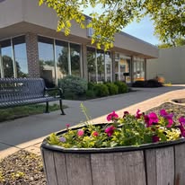 Flower bed with pink and purple flowers in front of a building with large windows and a bench nearby.