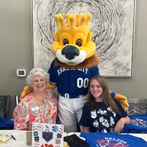 Person in lion mascot costume with two women, one older and one younger, sitting at a table with sports-themed items, in front of abstract artwork.