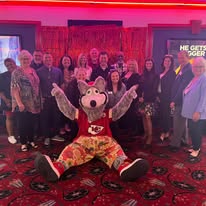 Group of people with a large mascot character in front, all smiling indoors with red lighting and patterned carpet.