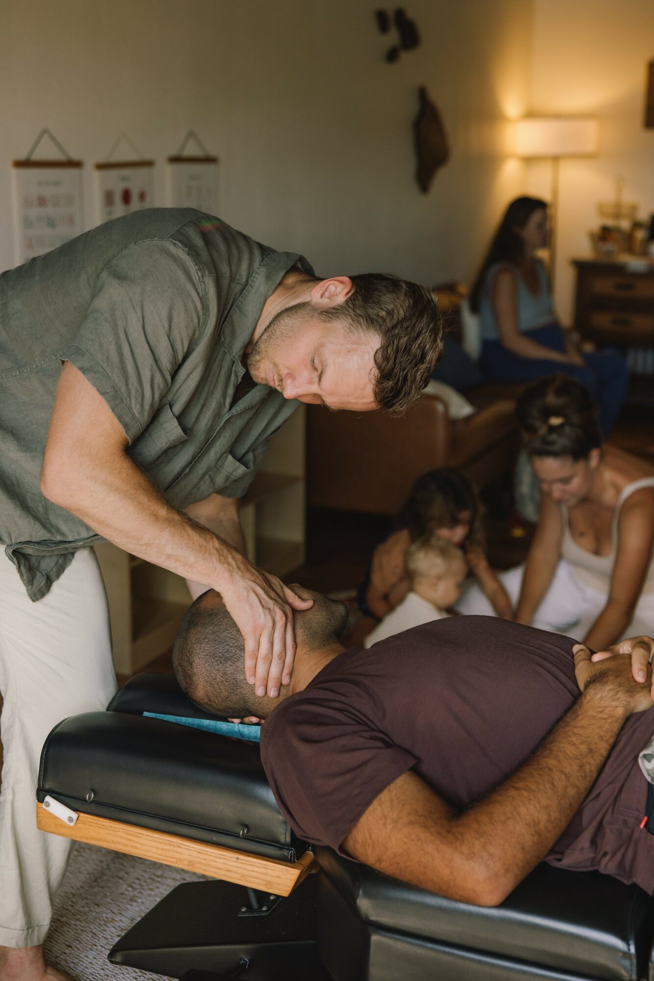 Man performing CPR on a person lying on a table, with children and a woman in the background in a room.