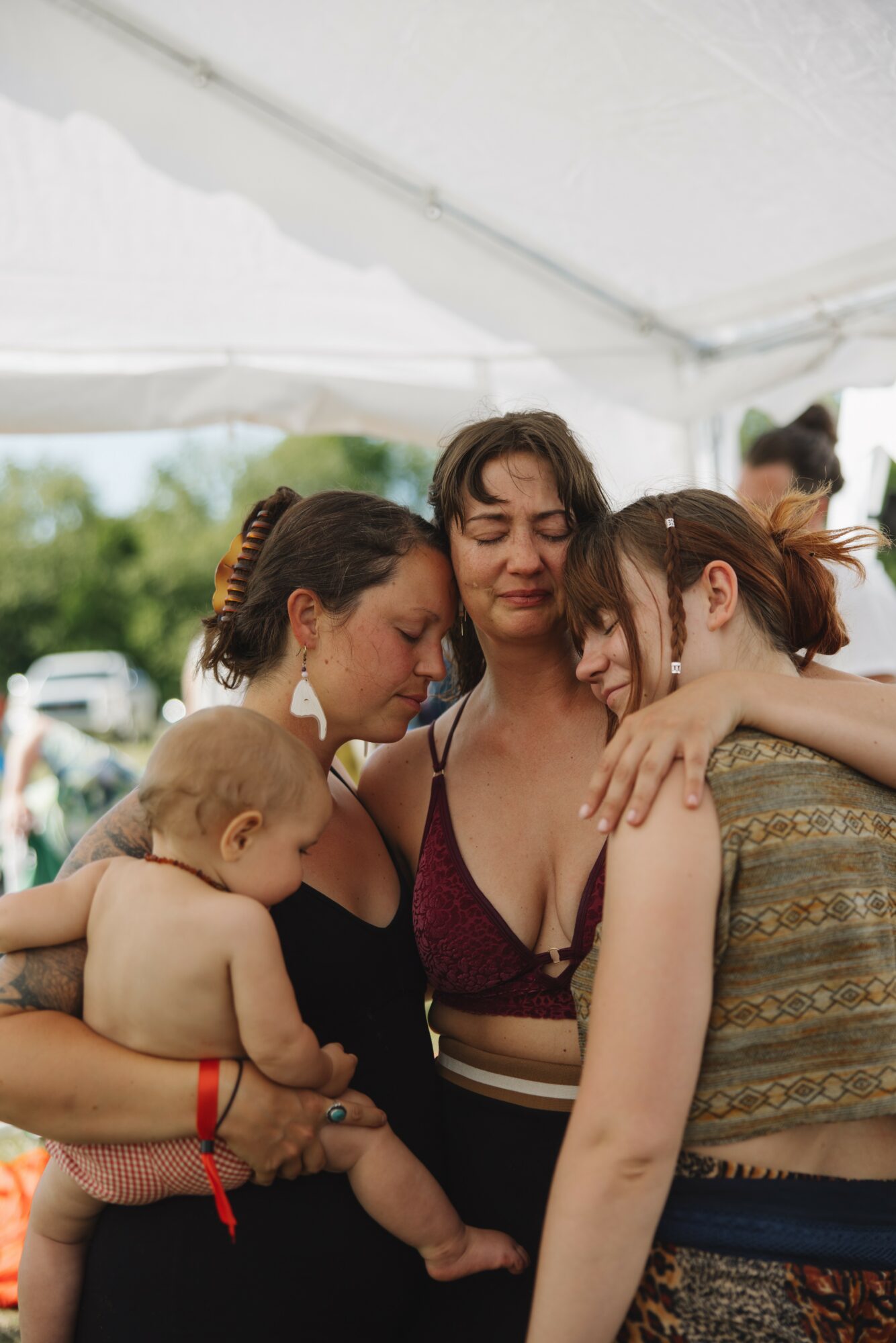 Four women and a baby under a white tent outdoors, embracing and talking, with trees and sky in background.