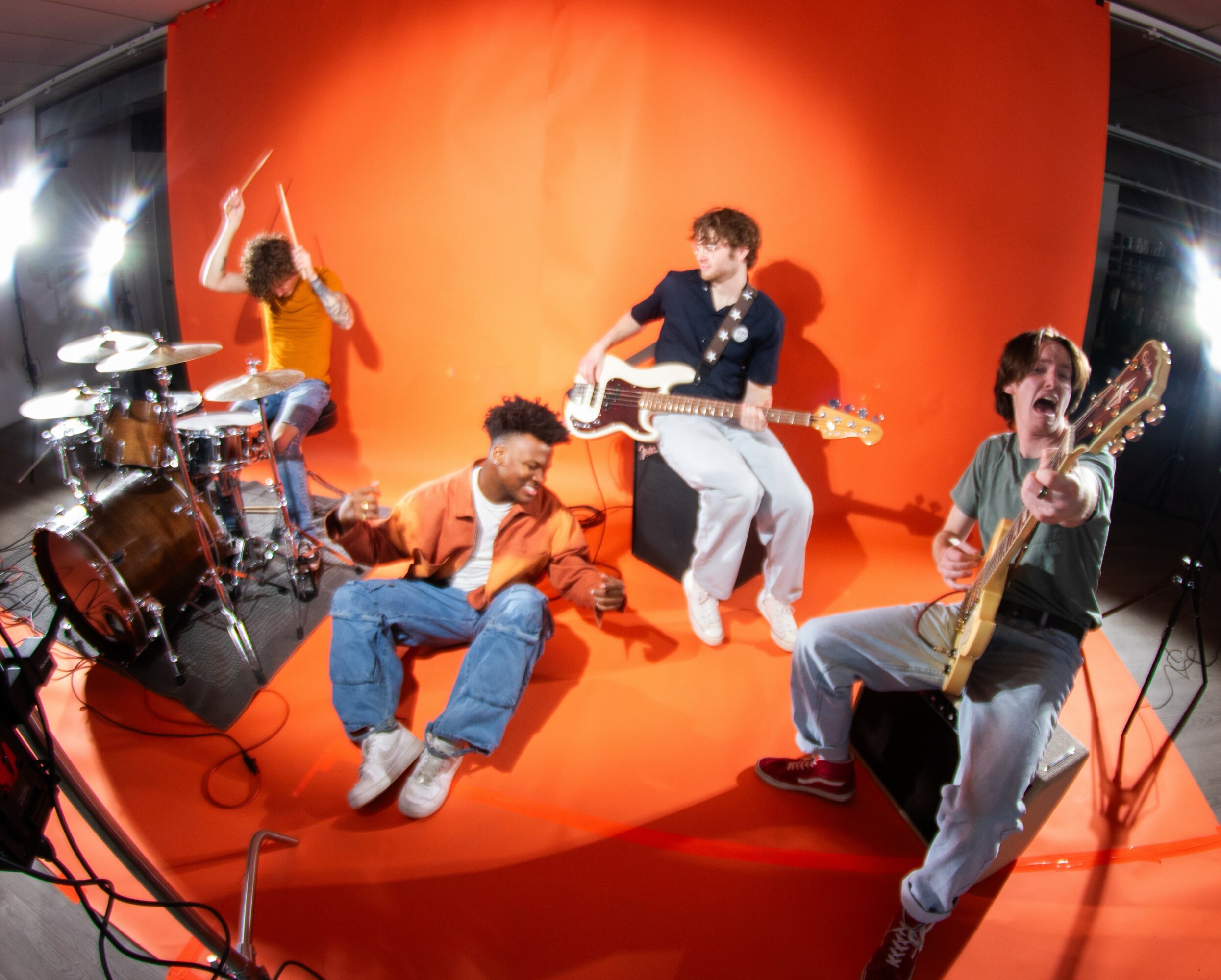 Four musicians perform on a red stage with a red backdrop, playing drums, guitar, and singing, with bright stage lights.