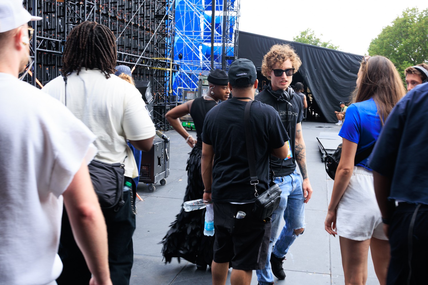 Group of people gathered outdoors near a stage with blue lighting, some wearing sunglasses and casual clothing.
