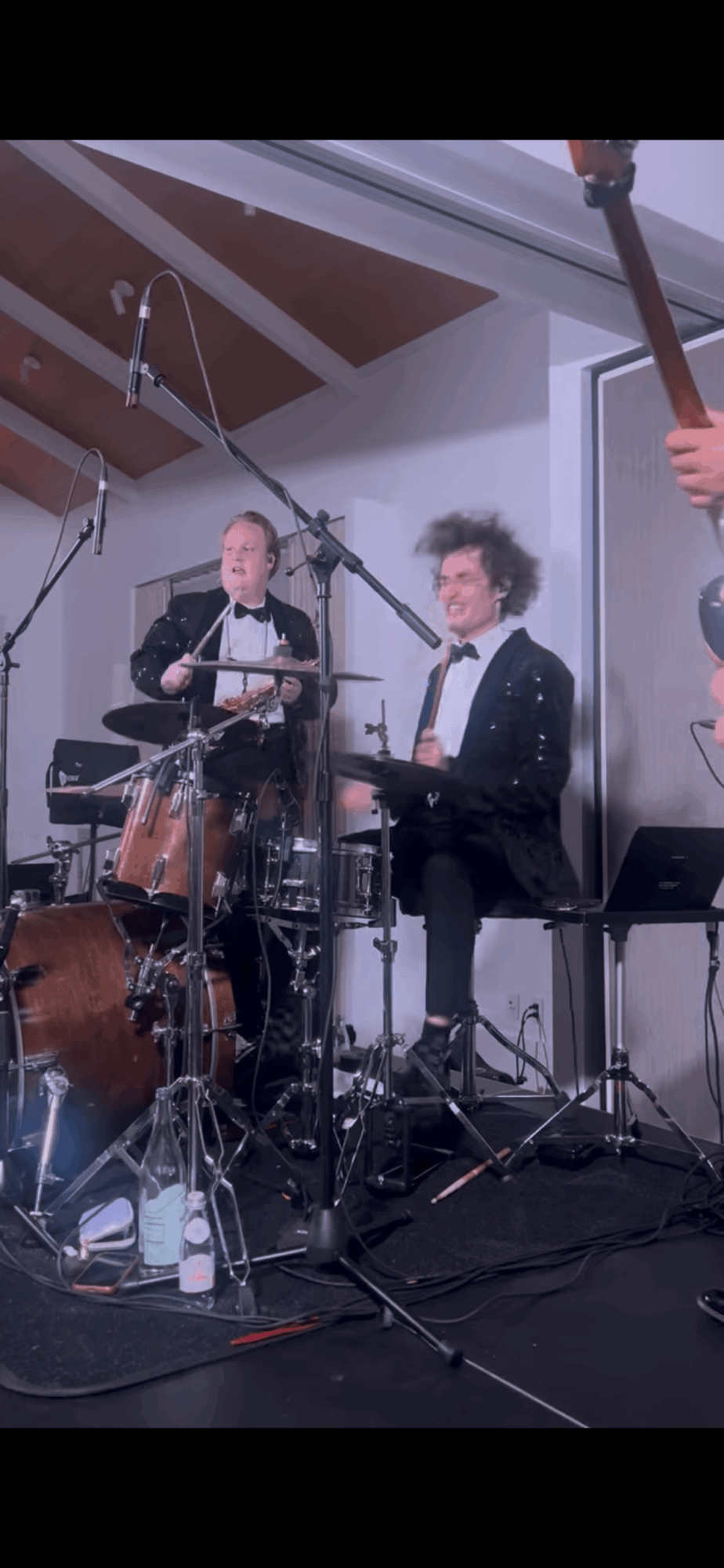 Two musicians perform on stage with drums and keyboard, one with curly hair, in a room with white walls and ceiling.