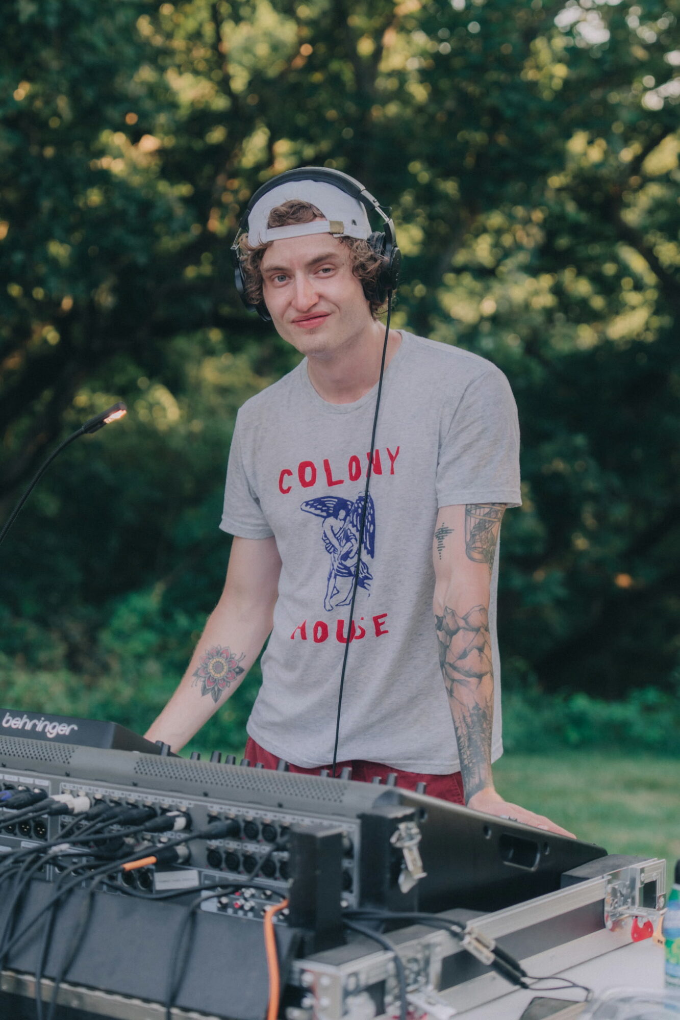 Young person with headphones and tattoos standing outdoors near a DJ equipment, trees in background.