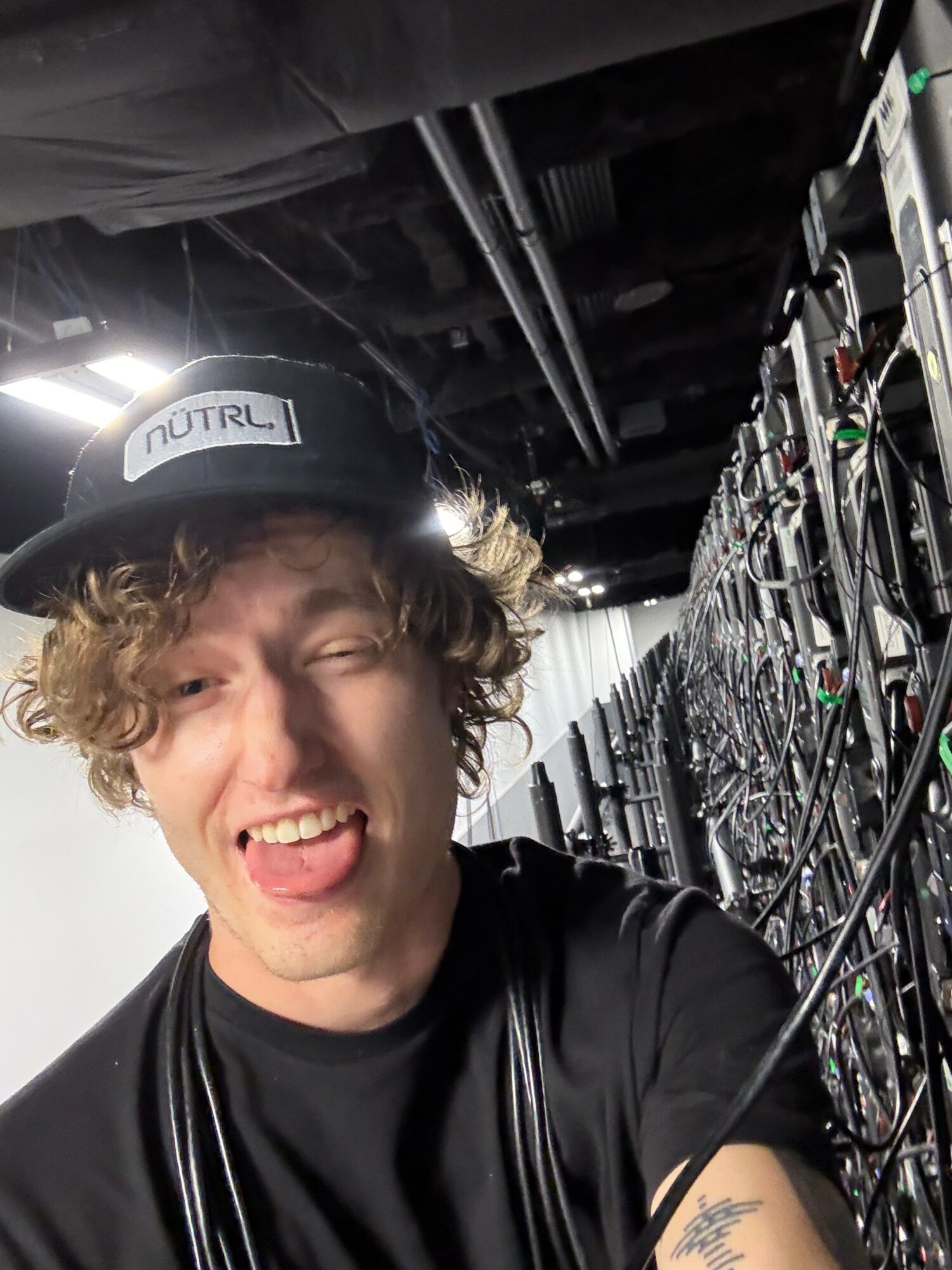 Young person with curly hair wearing a cap, smiling and sticking out tongue, in a room with electronic equipment.