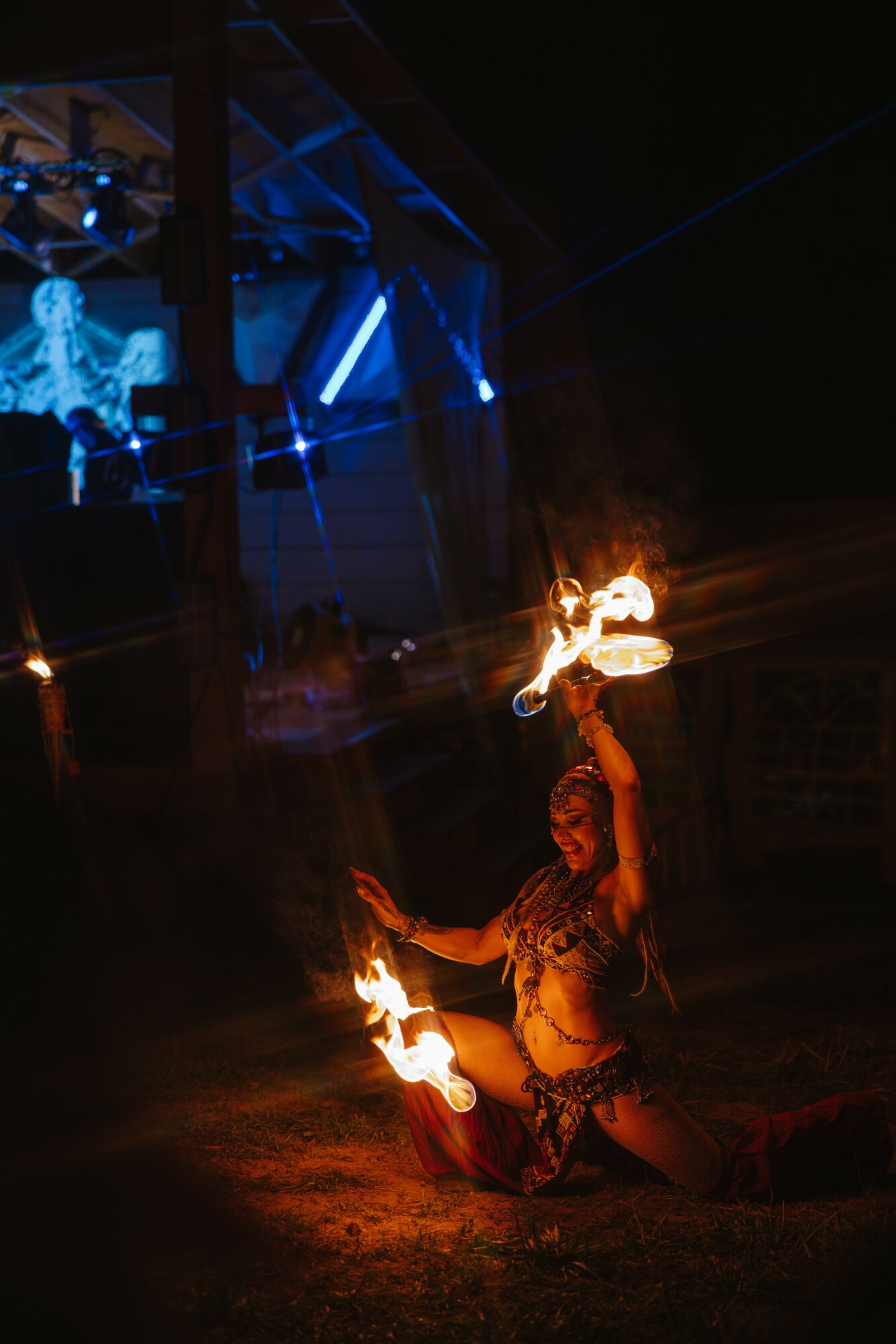 A performer with fire props kneels on the ground at night, with a stage and a blue-lit skeleton figure in the background.