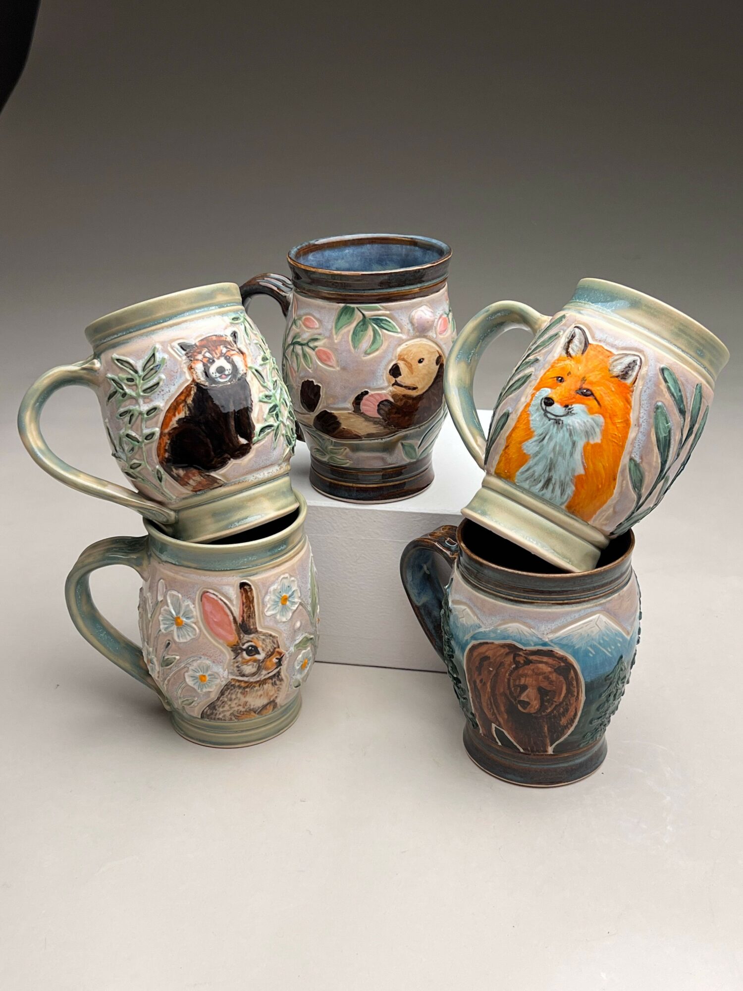 Four ceramic mugs with animal and nature designs, stacked and leaning against a wall, on a white surface.