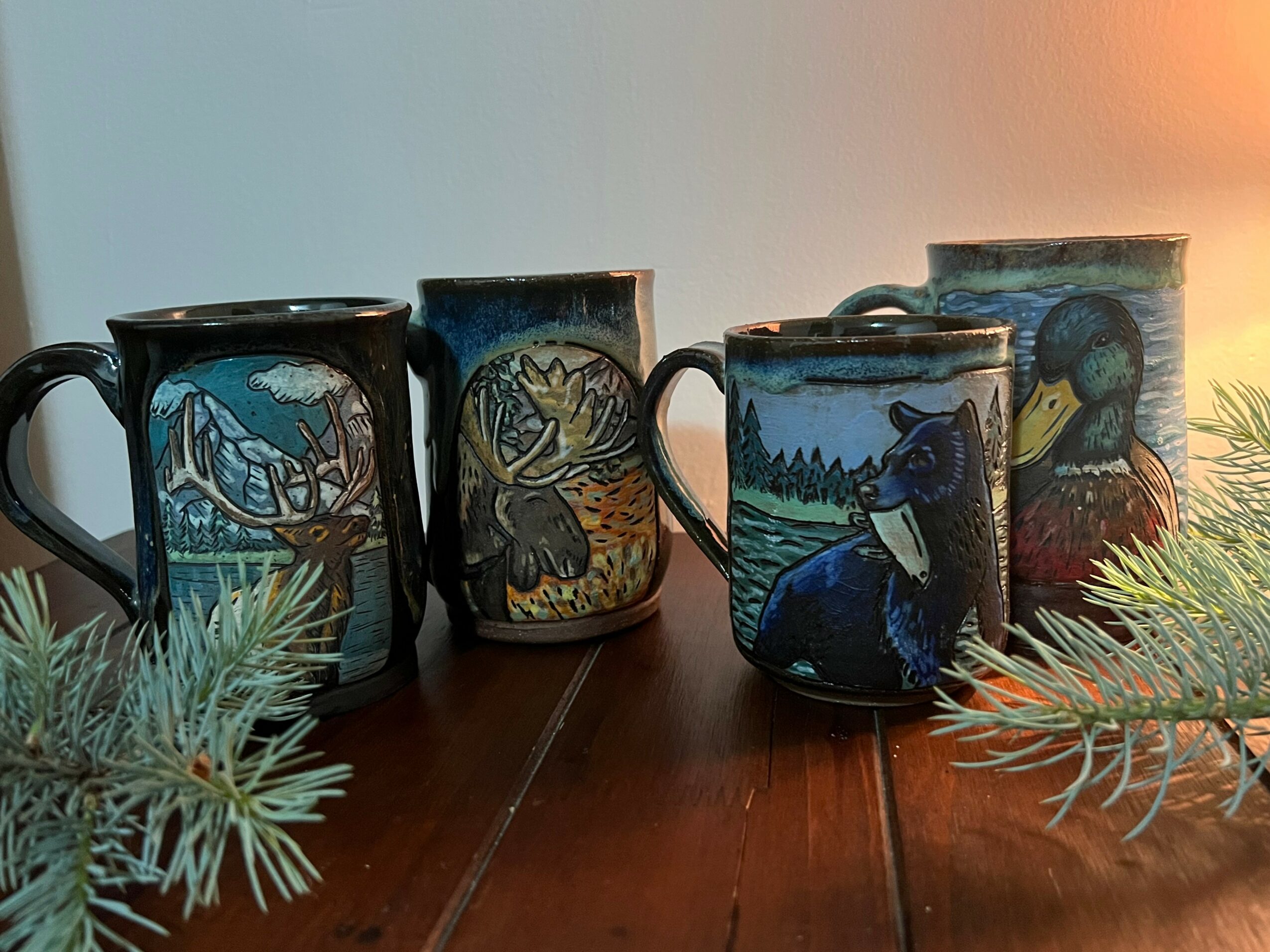 Five ceramic mugs with animal and nature designs on a wooden surface, with pine branches in the foreground.