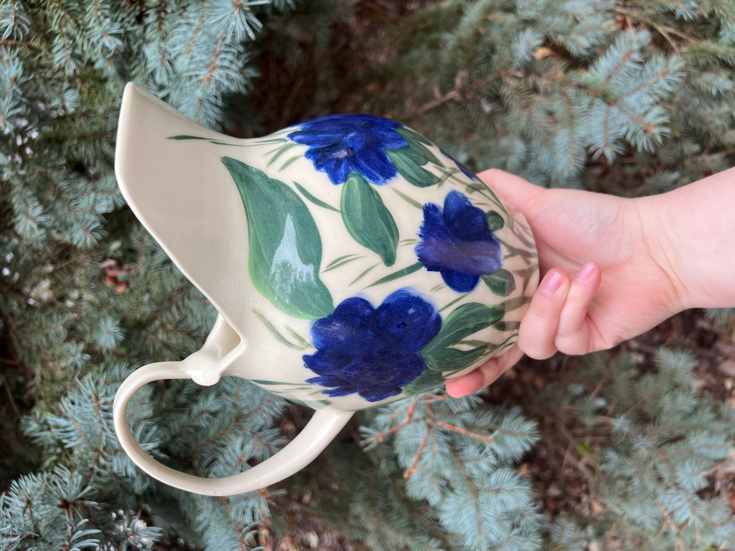 Person holding a floral-patterned teapot with blue flowers and green leaves, against a background of pine branches.