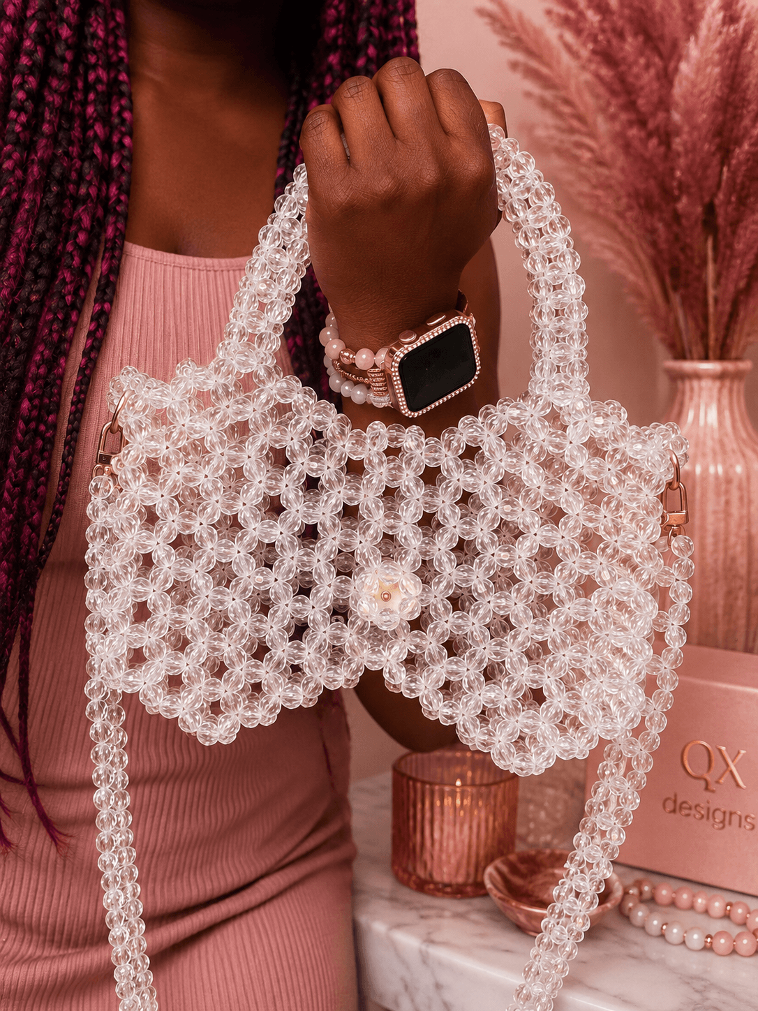 Person holding a white, beaded handbag with a pearl detail, wearing a smartwatch and pink clothing, with pink decor in background.