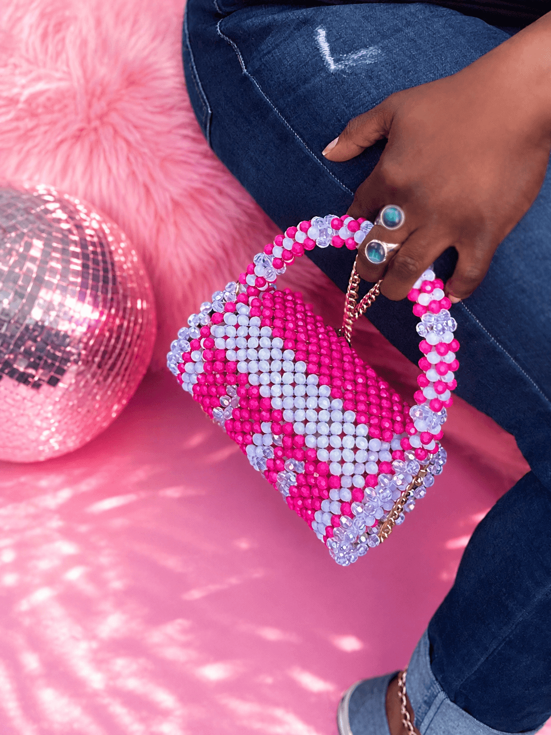 Person holding a pink and white beaded handbag with rings on fingers, sitting on pink fluffy surface with a disco ball nearby.