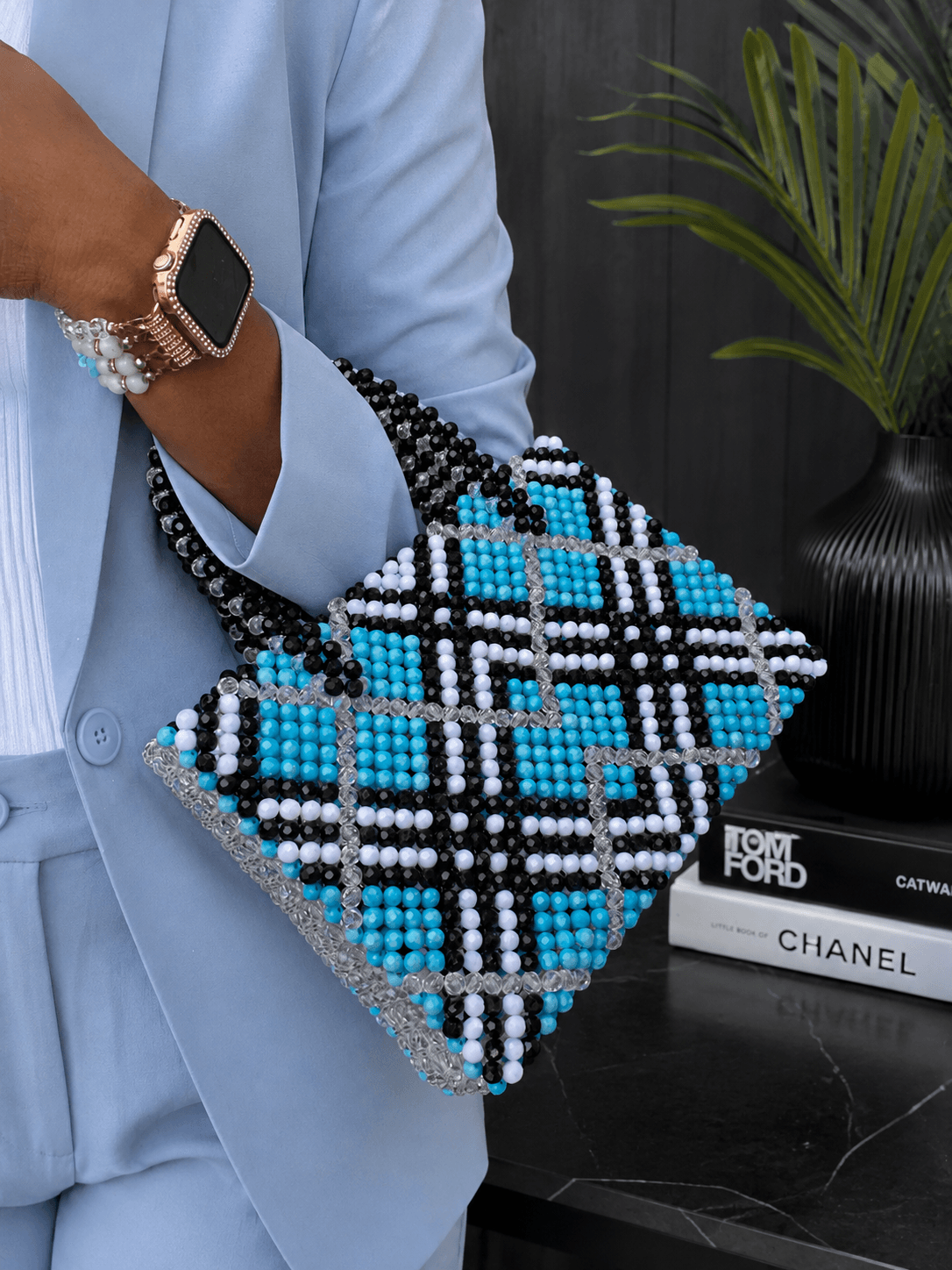 Person holding a beaded handbag with black, white, and blue beads, wearing a light blue shirt and a smartwatch, standing near a black table with books and a potted plant.