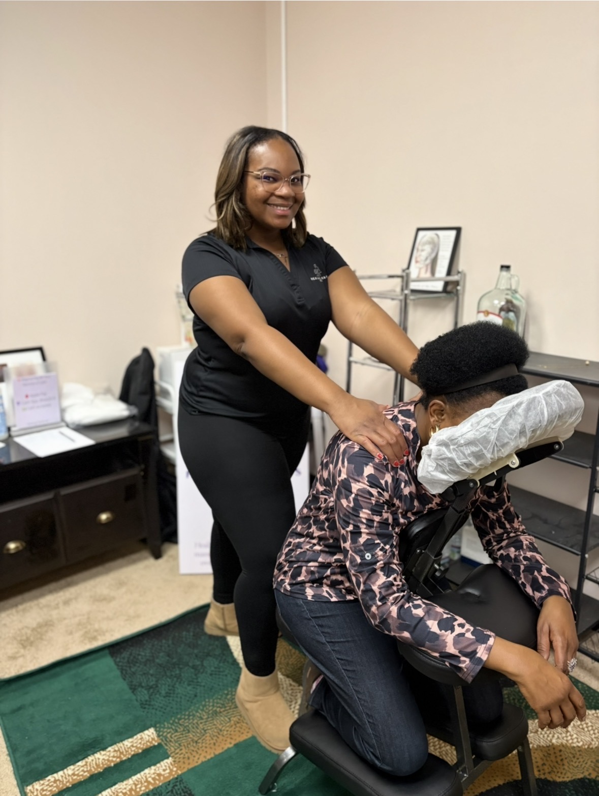 A woman standing and smiling while giving a shoulder massage to a seated person in a room with furniture and decorations.