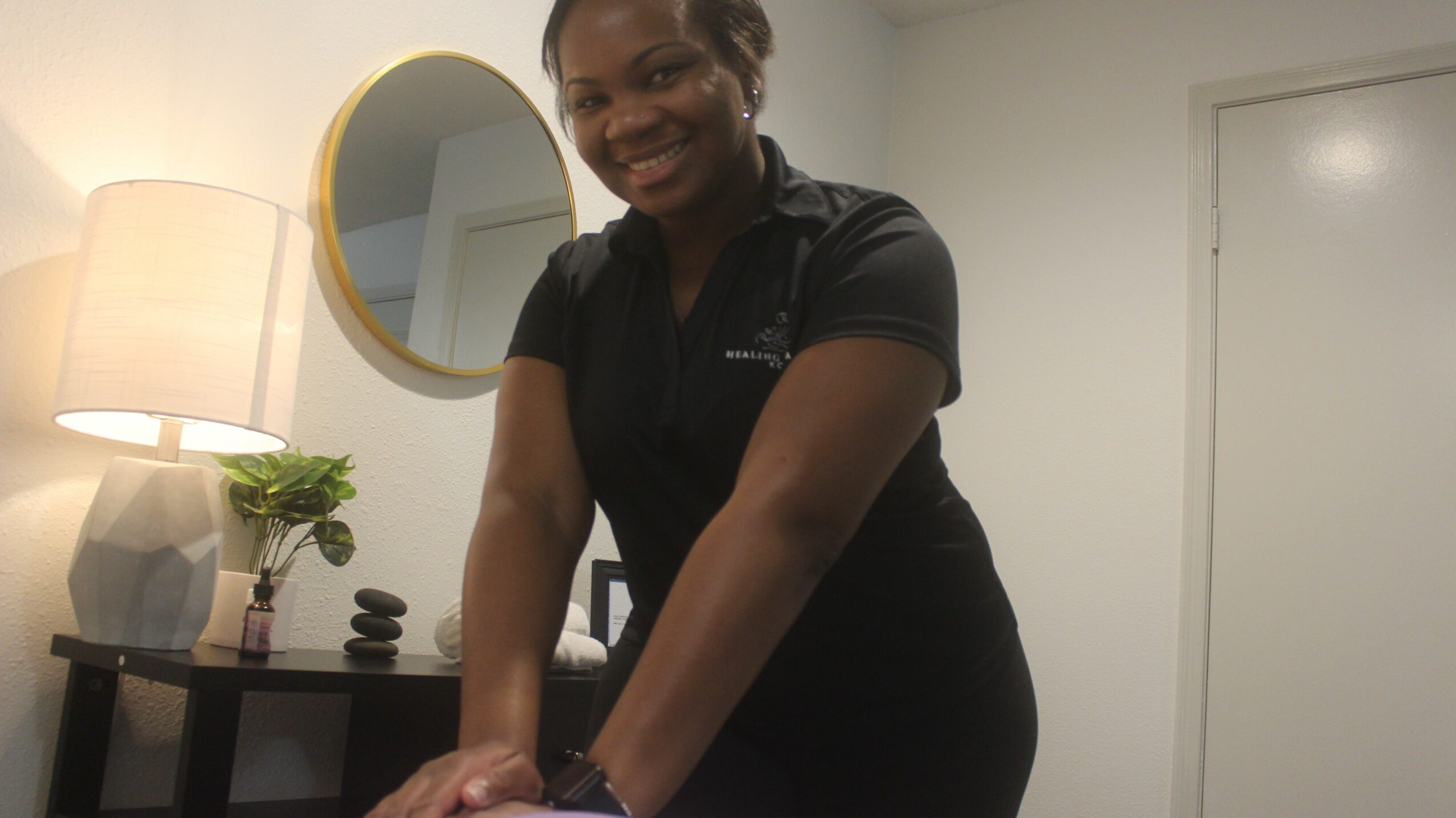 Smiling woman in black uniform leaning on a table in a room with a lamp and mirror.