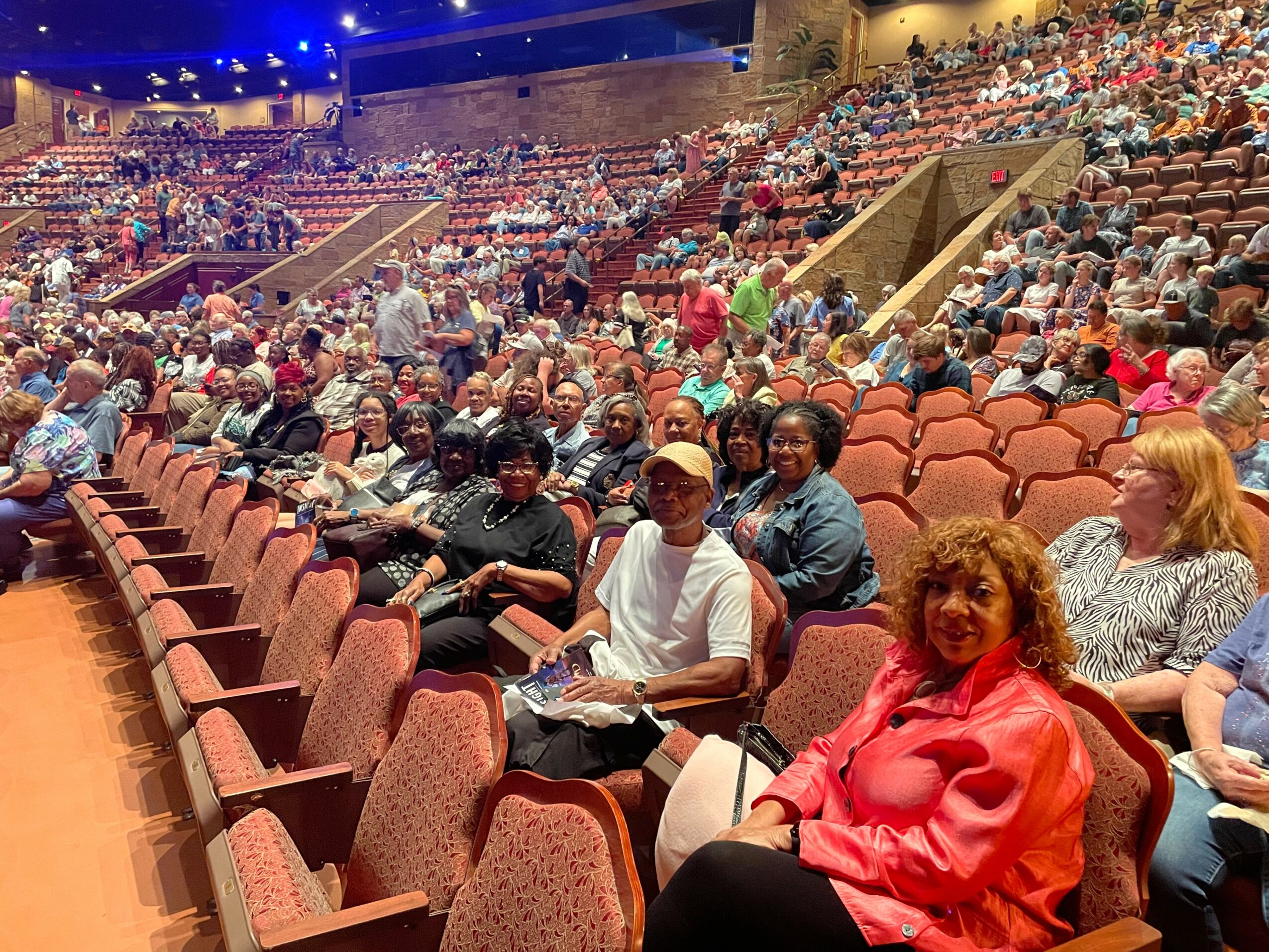 Audience seated in a large auditorium with many empty seats, some people standing and walking around, stage area in background.