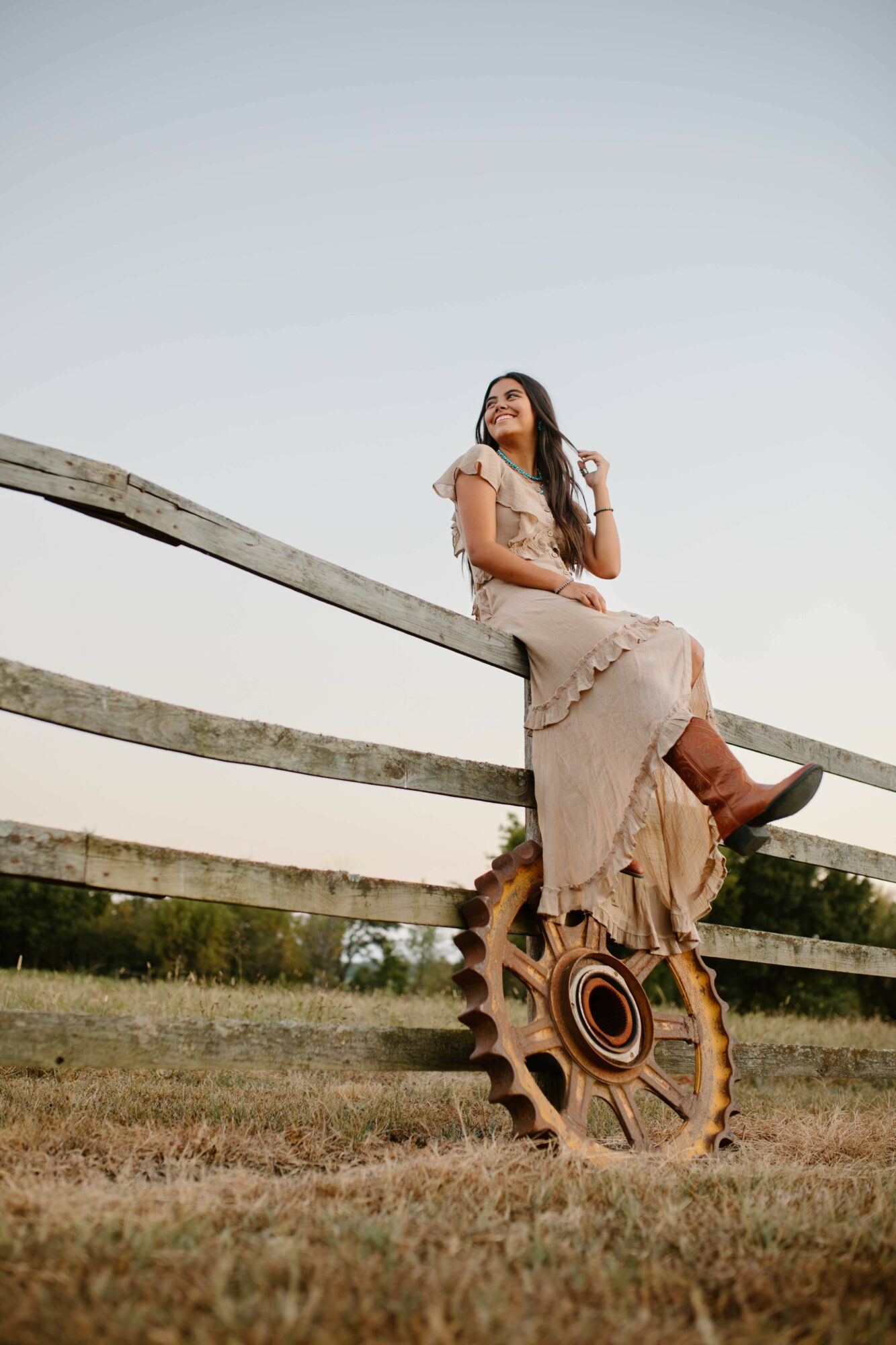 Woman sitting on a wooden fence with a gear wheel at the base, outdoors in a field, looking up, clear sky.