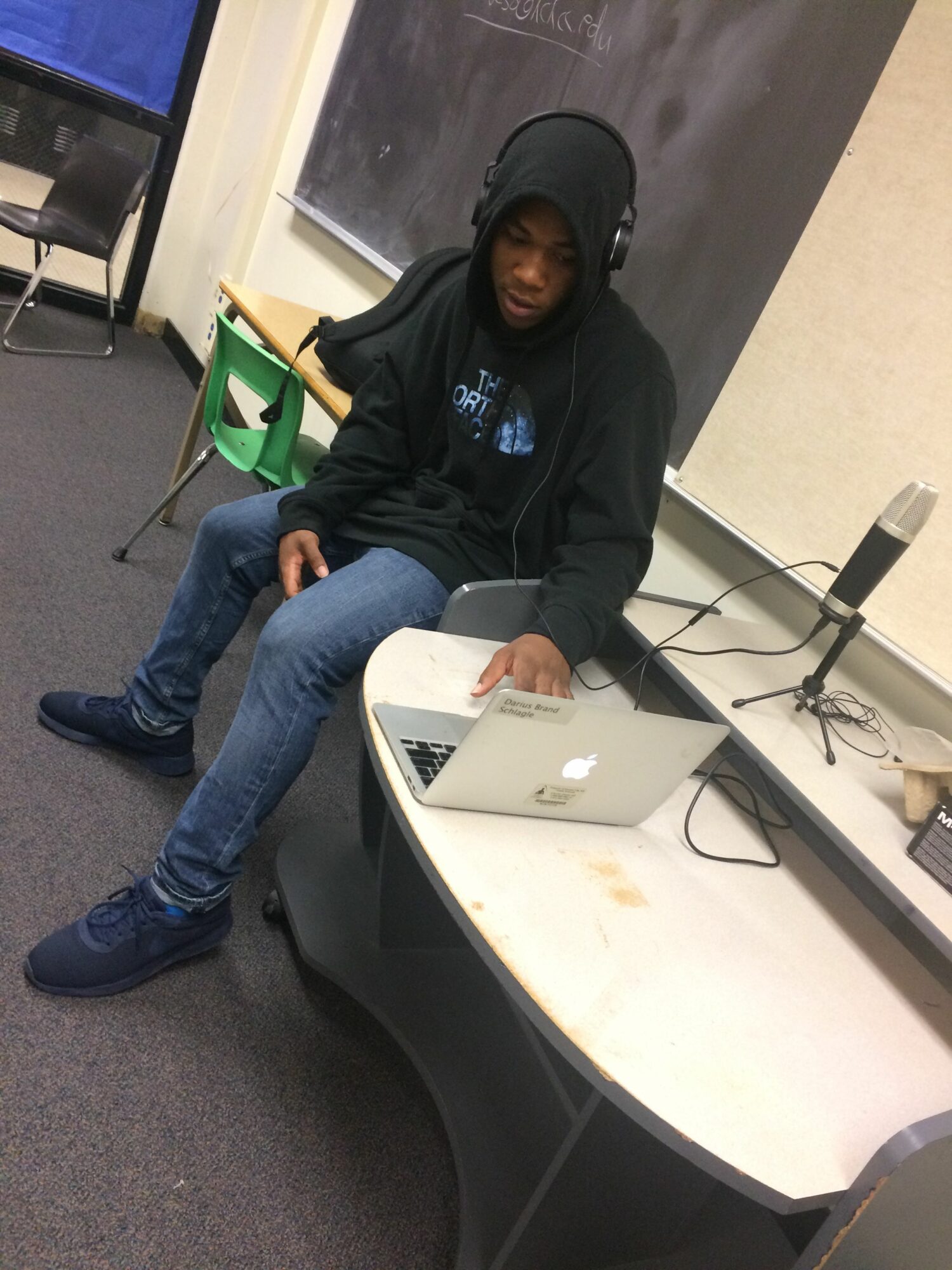 Person standing next to a desk with a laptop, in an office setting, wearing a black hoodie and jeans.