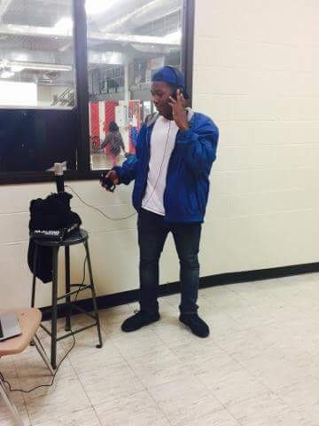 Person in blue jacket and cap standing indoors, holding headphones and a device, near a window and a stool.