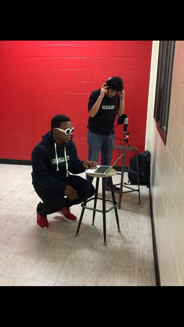 Two people in a room with a red wall, one sitting at a small table with a laptop, the other taking a photo.