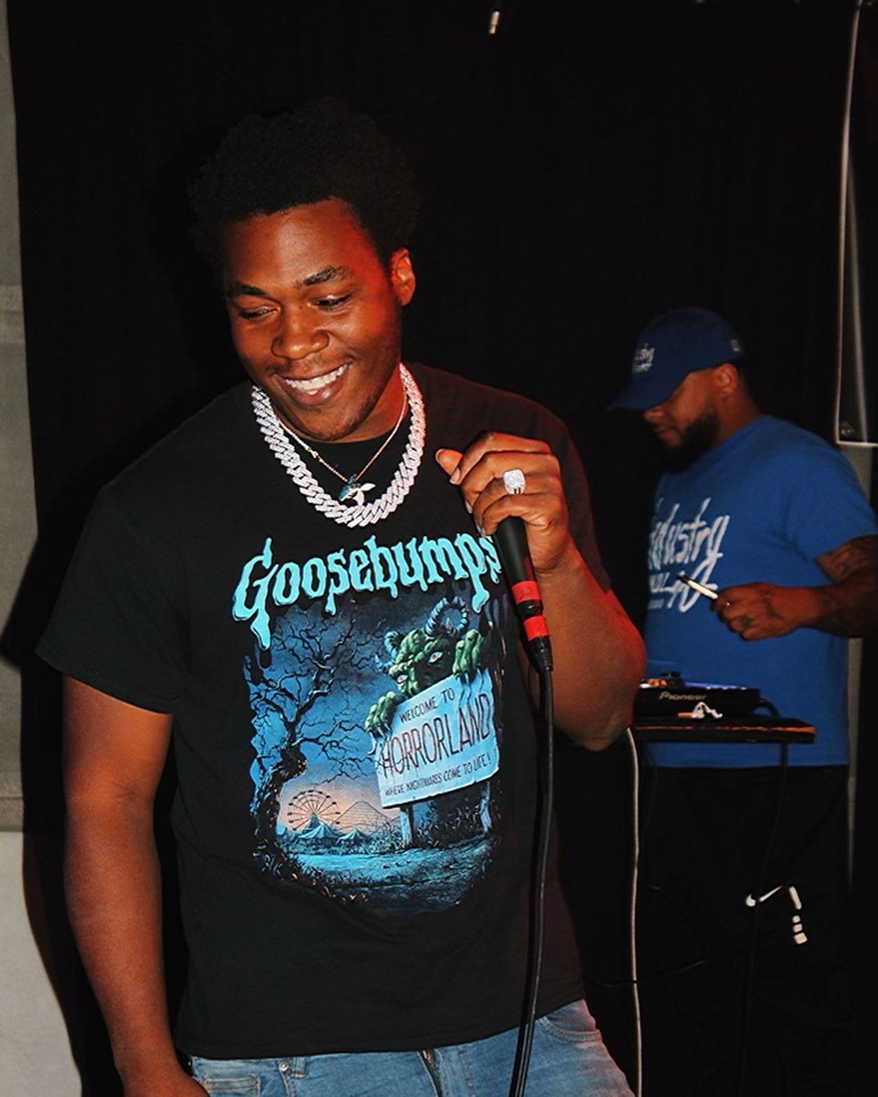 Person smiling, holding a microphone, wearing a black T-shirt with a graphic and text, with another person in the background.