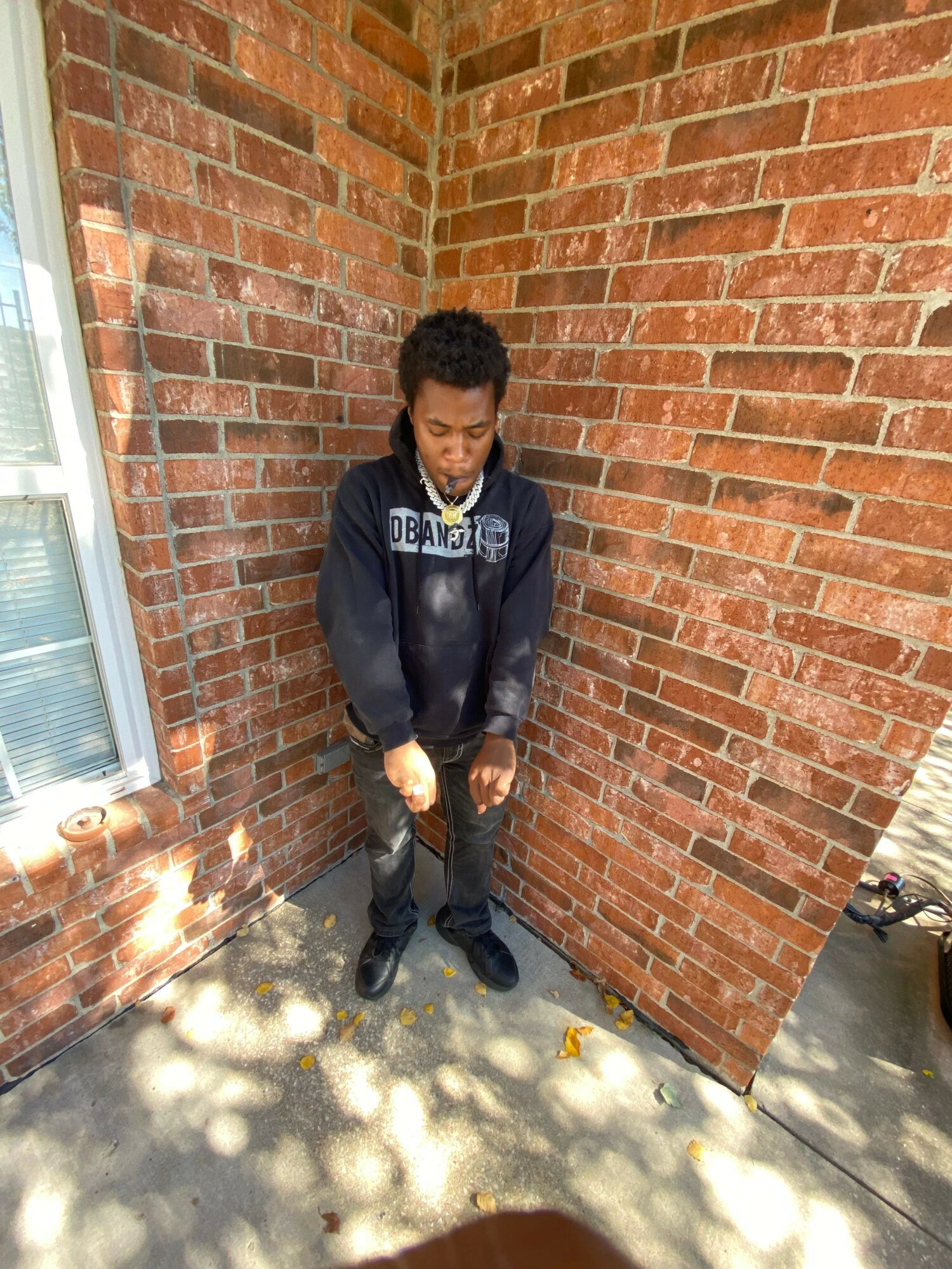 Person standing against a brick wall, wearing a dark jacket and pants, with sunlight and shadows on the ground.