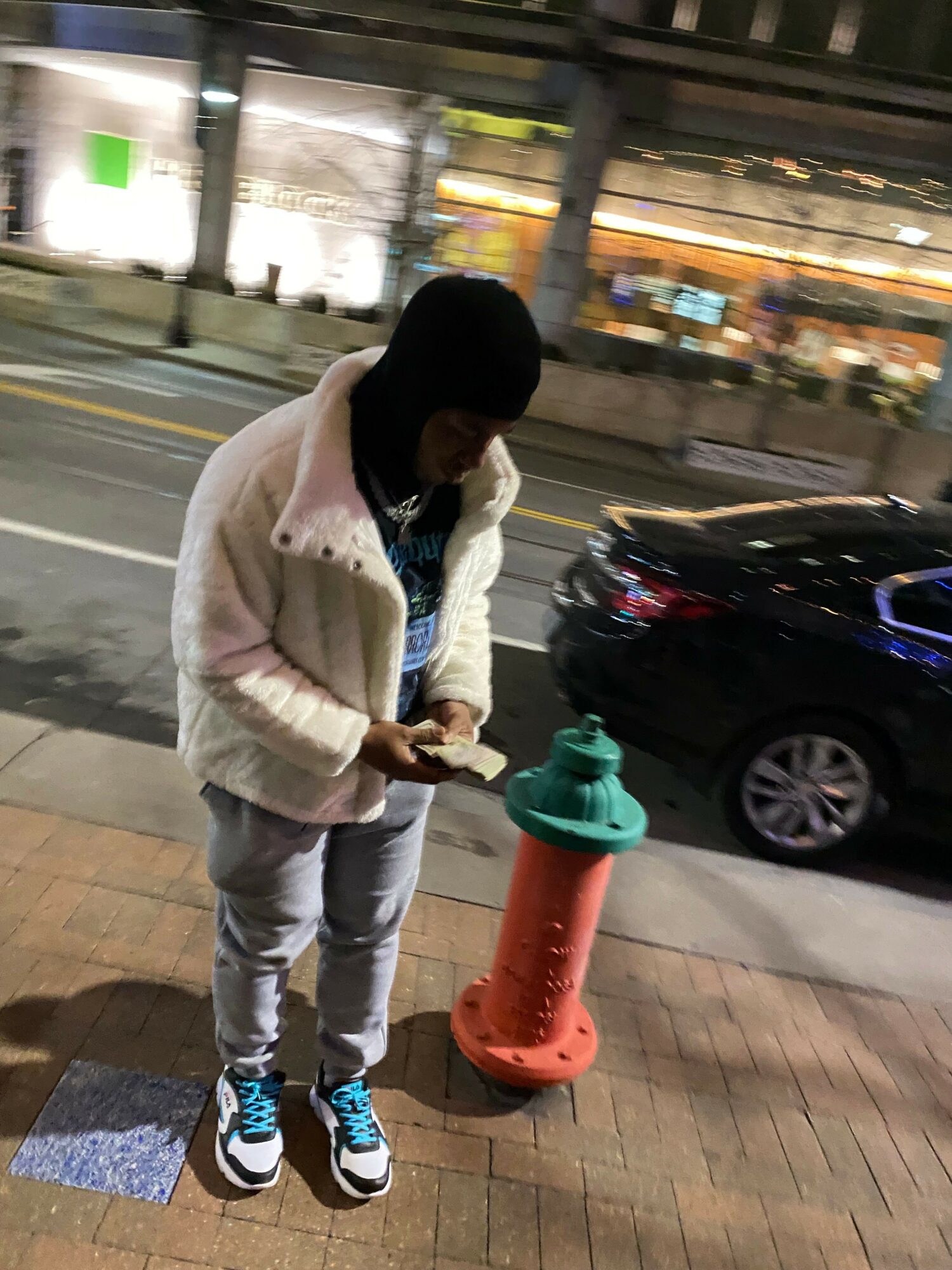 Person with long dark hair wearing a white jacket and gray pants standing on a sidewalk at night, holding a phone.