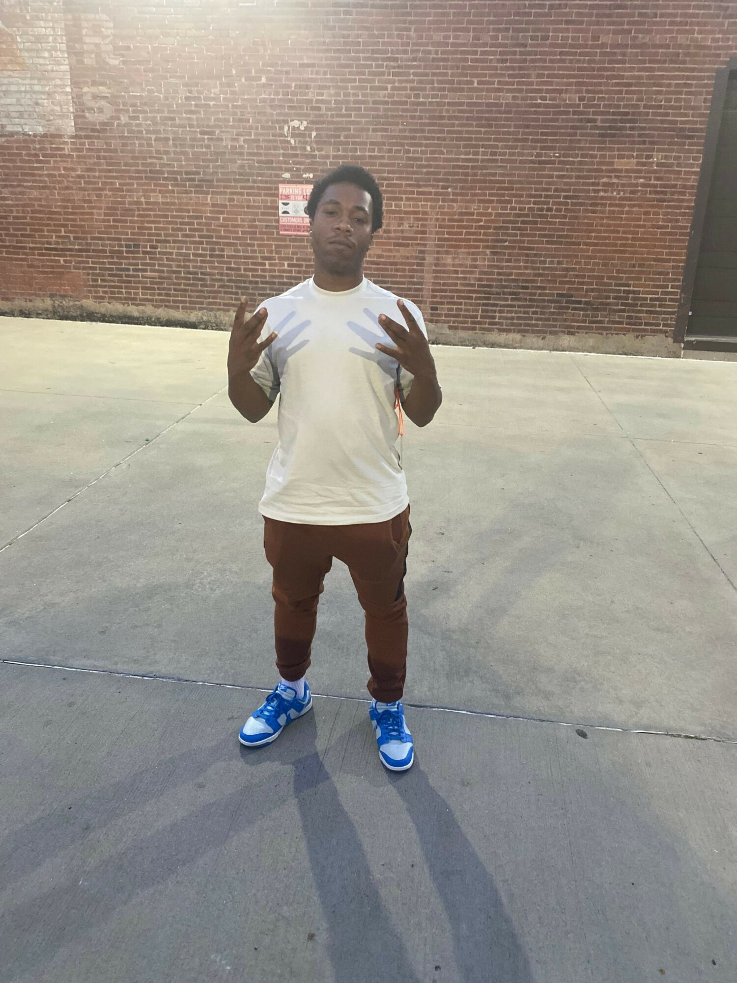 Person standing on concrete ground with a brick wall in the background, wearing a white shirt, dark pants, and blue shoes.