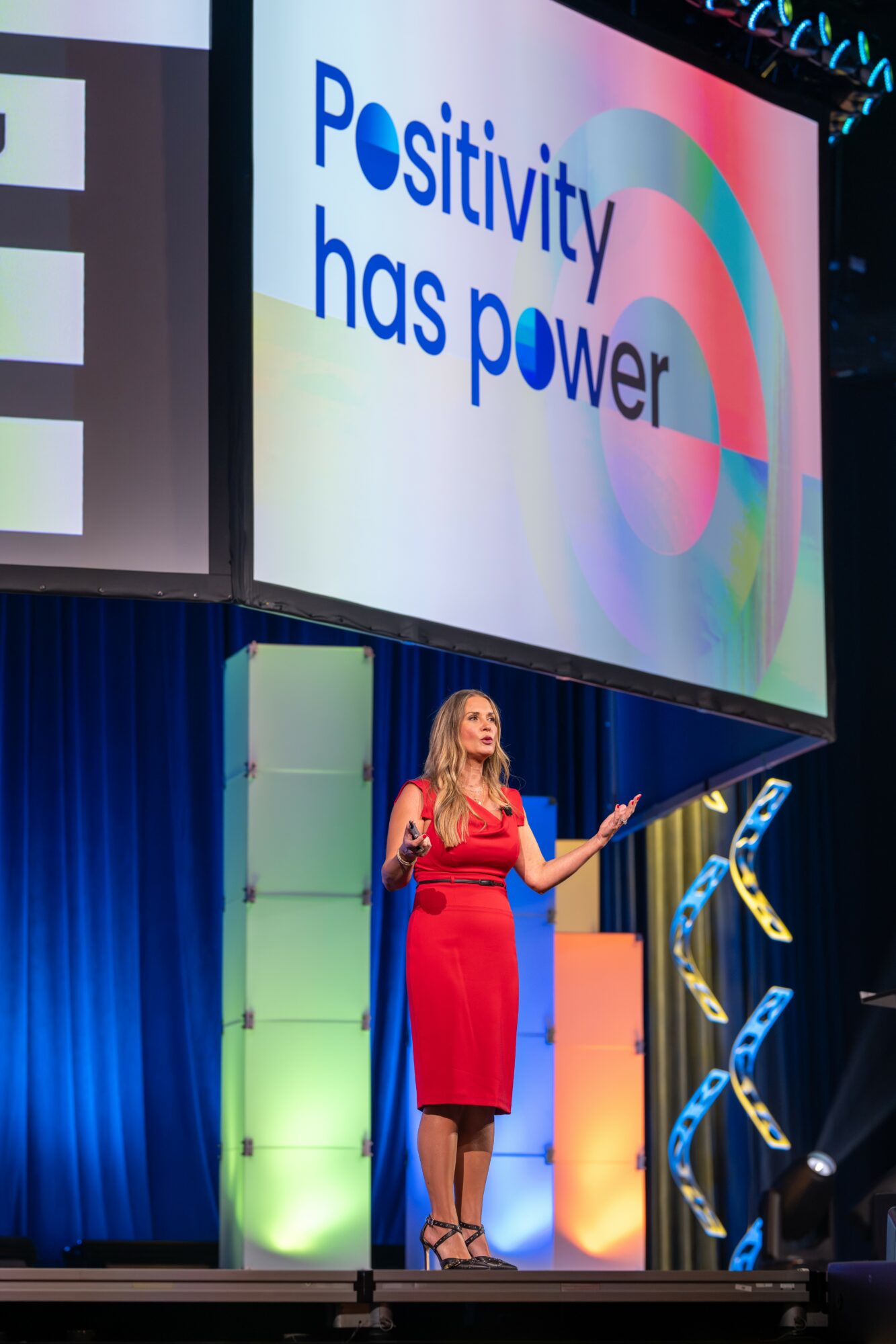 Woman in red dress speaking on stage with a large screen displaying 'Positivity has power' behind her.