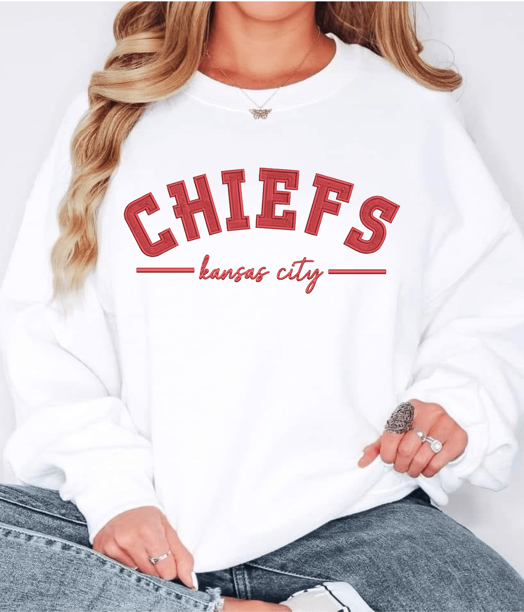 Person wearing a white sweatshirt with red text reading 'CHIEFS' and 'kansas city' underneath, with long wavy hair and rings on fingers.
