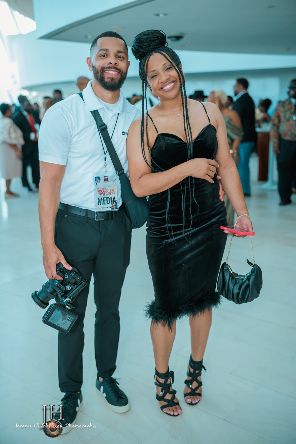 A man and woman smiling at an event, with the man holding a camera, in a bright indoor space with other people.