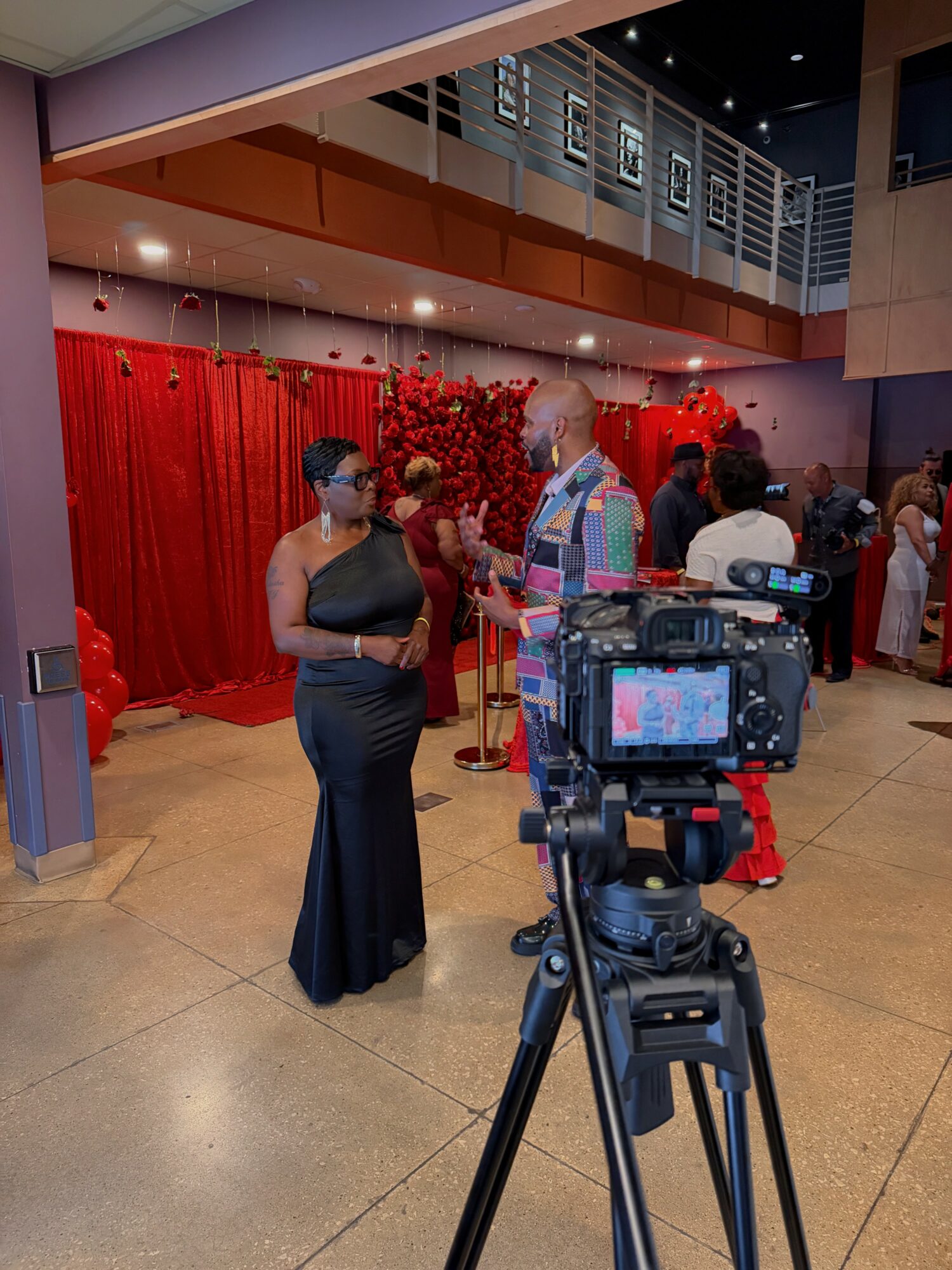 Two people in formal attire talking in front of a red curtain at an indoor event, with a camera on a tripod in the foreground.