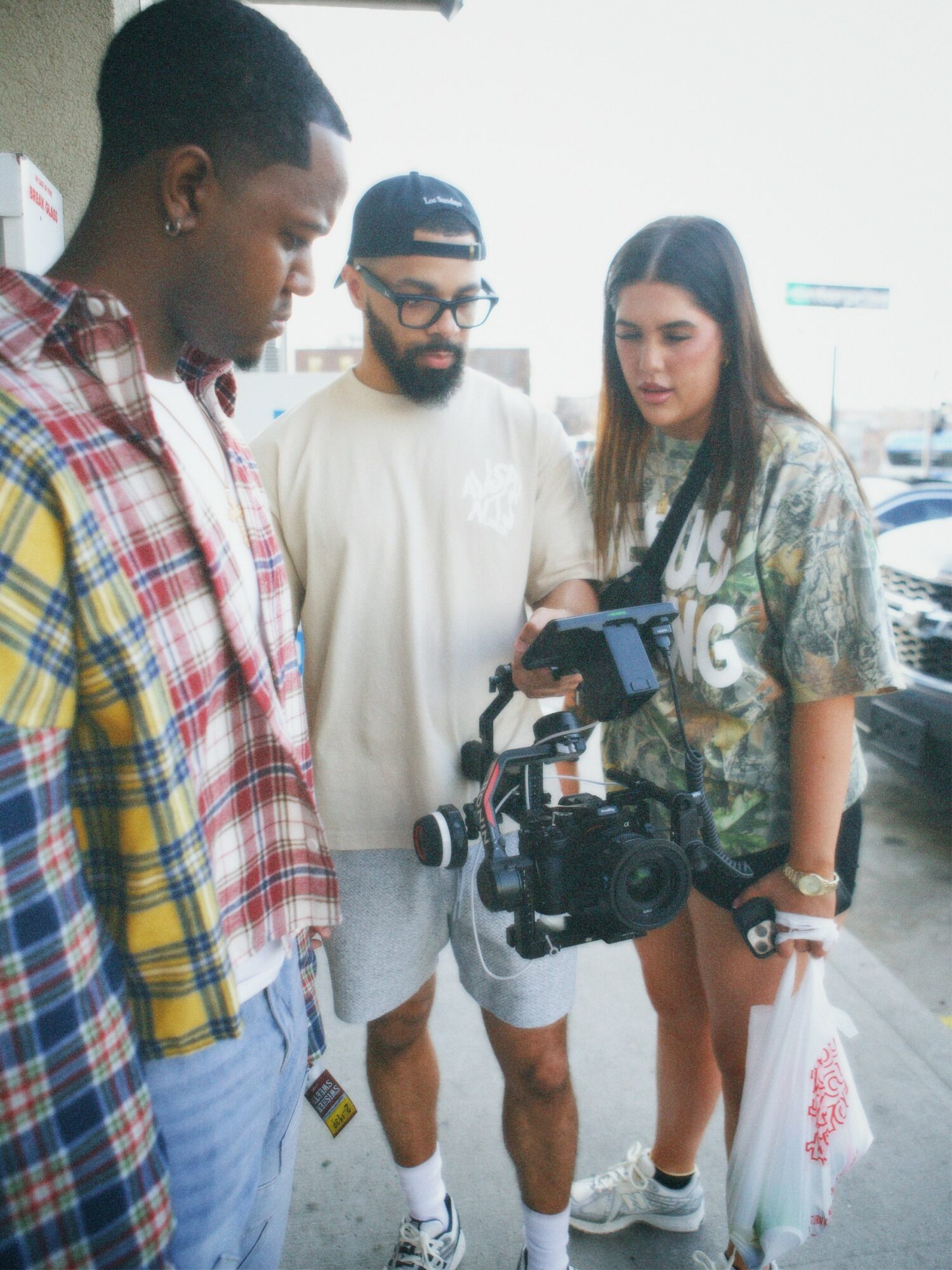 Three people look at a camera and a phone outside, with a bright background and parked cars visible behind them.