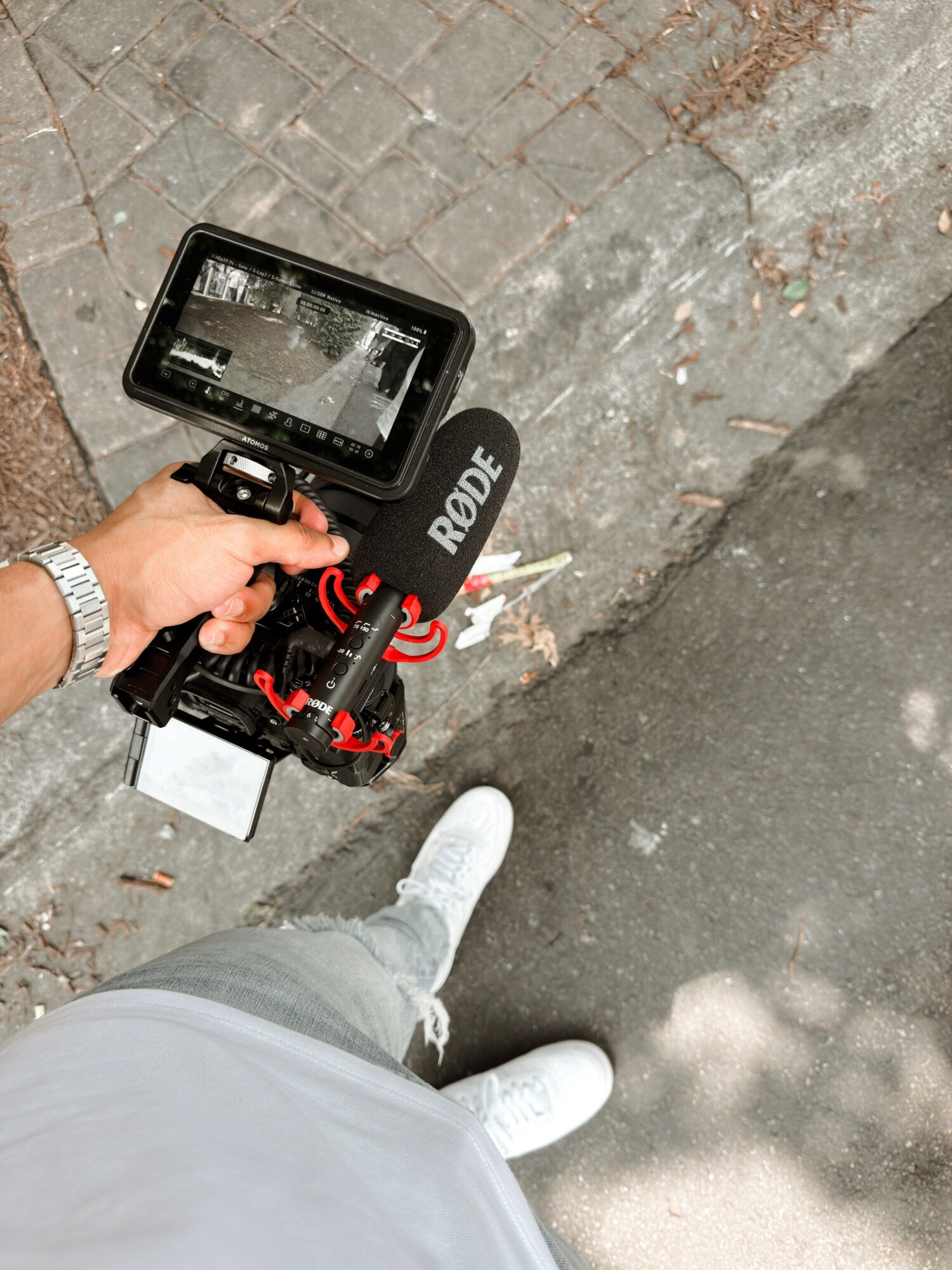 Person holding a camera with a Rode microphone, standing on a sidewalk with gray pavement and brick edge, wearing white shoes and light-colored pants.